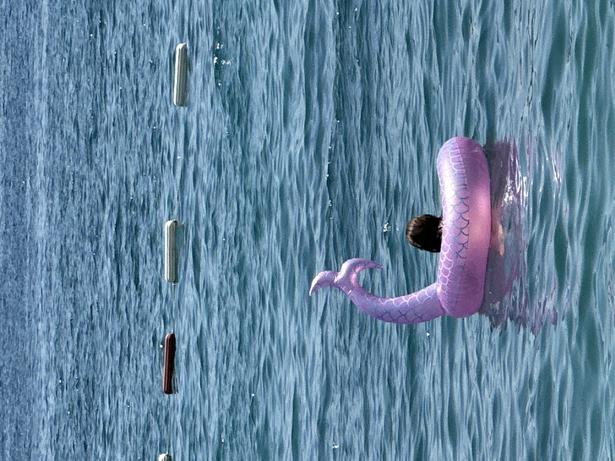 This media file depicts a person swimming in the sea near Budva, Montenegro. The water is a vibrant blue, with textured ripples suggesting a gentle current or waves. Floating vertically in the water, spaced apart, are several slender, elongated objects. One object is a dark brown, cylindrical shape, while two others appear to be metallic, possibly gray or silver.
The main subject is a person partially submerged in the water, wearing a pink, inflatable float shaped like a mermaid's tail. The float has a textured pattern of scales and a distinct fin at the end. The person's head and shoulders are visible, with dark hair. The activity taking place is swimming or floating in the water, with the mermaid tail float adding a playful element.
The scene appears to be outdoors during daylight, with the bright water and sunlight reflecting off its surface. The location is suggested to be Budva, Montenegro, a coastal town known for its beaches and clear waters. There is no visible text in the image. The overall mood is one of leisure and enjoyment of a sunny day by the sea.