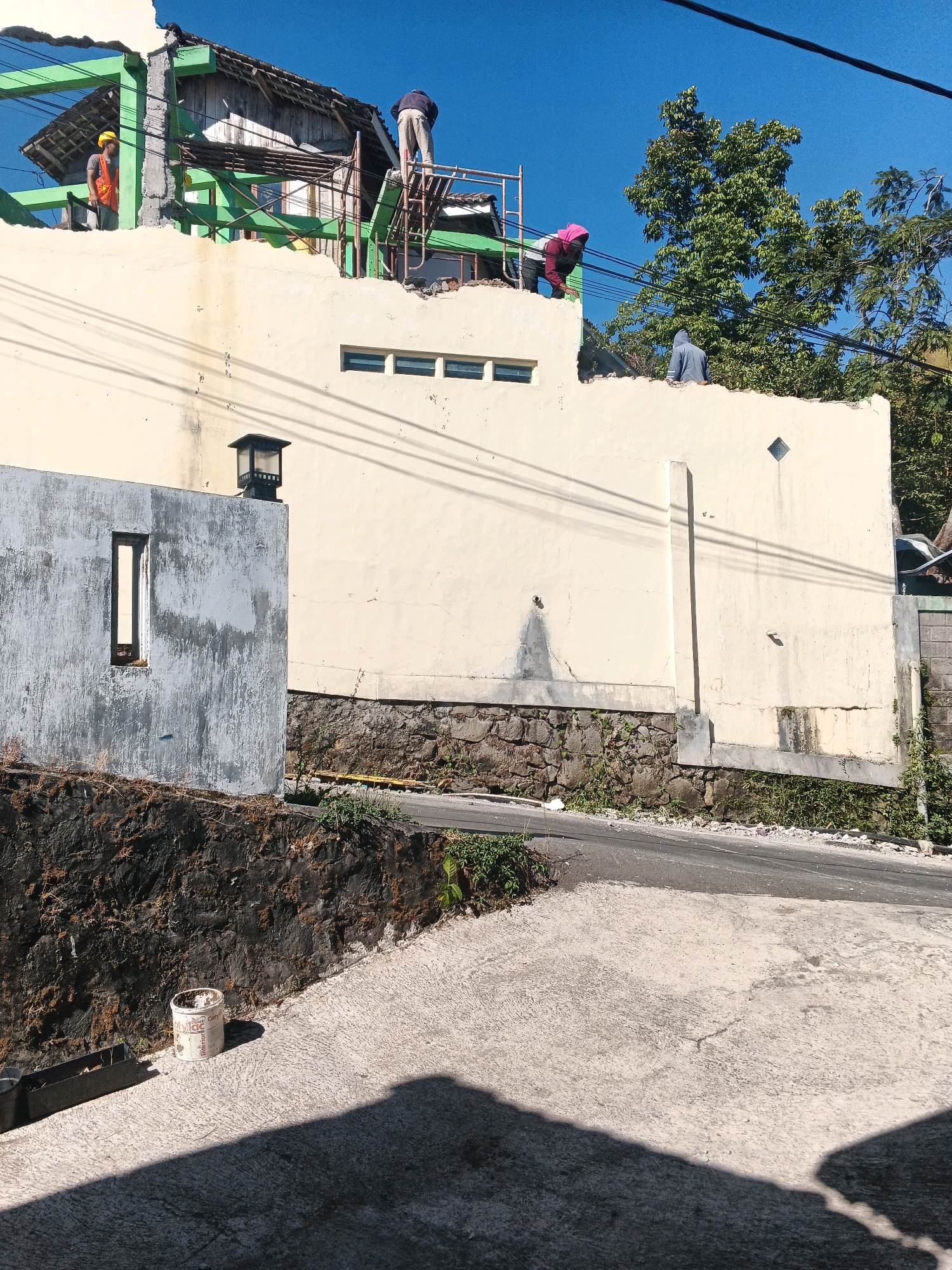 This image depicts a construction site in Keji, Indonesia, with multiple individuals engaged in work on a partially demolished building. In the foreground, a concrete driveway leads upwards, casting a shadow from an unseen object. A discarded white paint can and a metal trough are visible on the driveway. A stone retaining wall borders the driveway, supporting a grassy area.
The main focus is on the damaged building, which features a light yellow exterior wall with several small, rectangular windows. The roof appears to be undergoing reconstruction or demolition, with exposed wooden beams and roofing materials.
Three construction workers are visible on or near the damaged roofline. One worker, wearing a yellow hard hat and an orange vest, stands on scaffolding. Another worker, wearing a pink headscarf, is leaning over the wall, appearing to be working. A third worker, in a gray hooded jacket, is standing on the roof.
The background shows lush green trees and a clear blue sky, indicating an outdoor setting. Electrical wires are strung across the scene, and a black outdoor lamp is mounted on a separate wall structure to the left of the main building. The overall impression is one of ongoing construction or repair work in a rural or semi-urban environment.