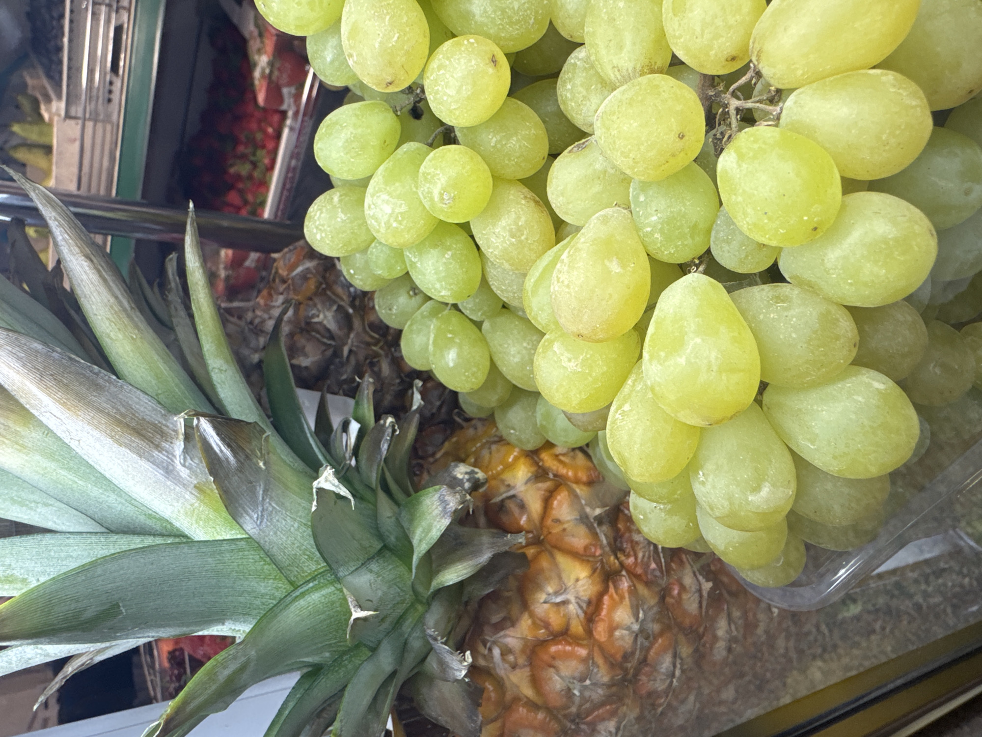 The image shows a pineapple and a bunch of green grapes in a plastic container, likely for sale. The fruit is displayed in an unidentified location in Moscow, Russia. The background reveals metal shelving and glimpses of other produce, including what appears to be strawberries. The focus is on the presentation of the pineapple and grapes, suggesting a grocery store or market setting. No people are visible in the frame.