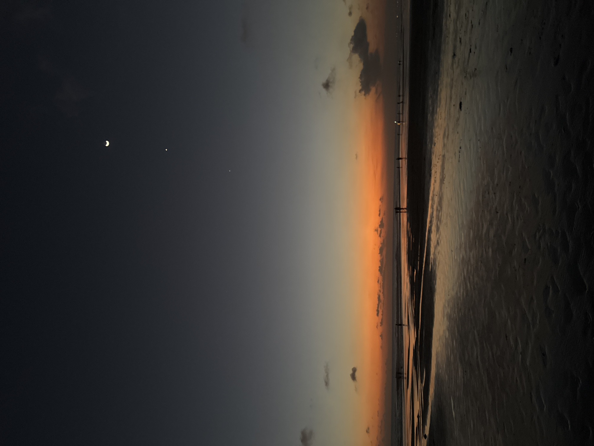 The image depicts a serene beach scene at dusk in Kuta, Indonesia. The sky transitions from a deep twilight blue on the left to a warm, vibrant orange near the horizon, indicating the sun has recently set. A crescent moon and a bright star are visible in the darkening sky, adding to the celestial display.
Along the horizon, a line of silhouetted figures can be seen standing on the wet sand near the water's edge. They appear to be enjoying the end of the day. The wet sand reflects the soft light of the sky, creating a shimmering effect. Further to the right, the beach stretches out, its surface textured with the marks of receding waves and possibly footprints, appearing dark in the fading light.
The overall mood is peaceful and contemplative, capturing the tranquil beauty of a tropical beach as day gives way to night. The scene suggests a casual stroll or moment of reflection by the sea. There is no visible text in the image.