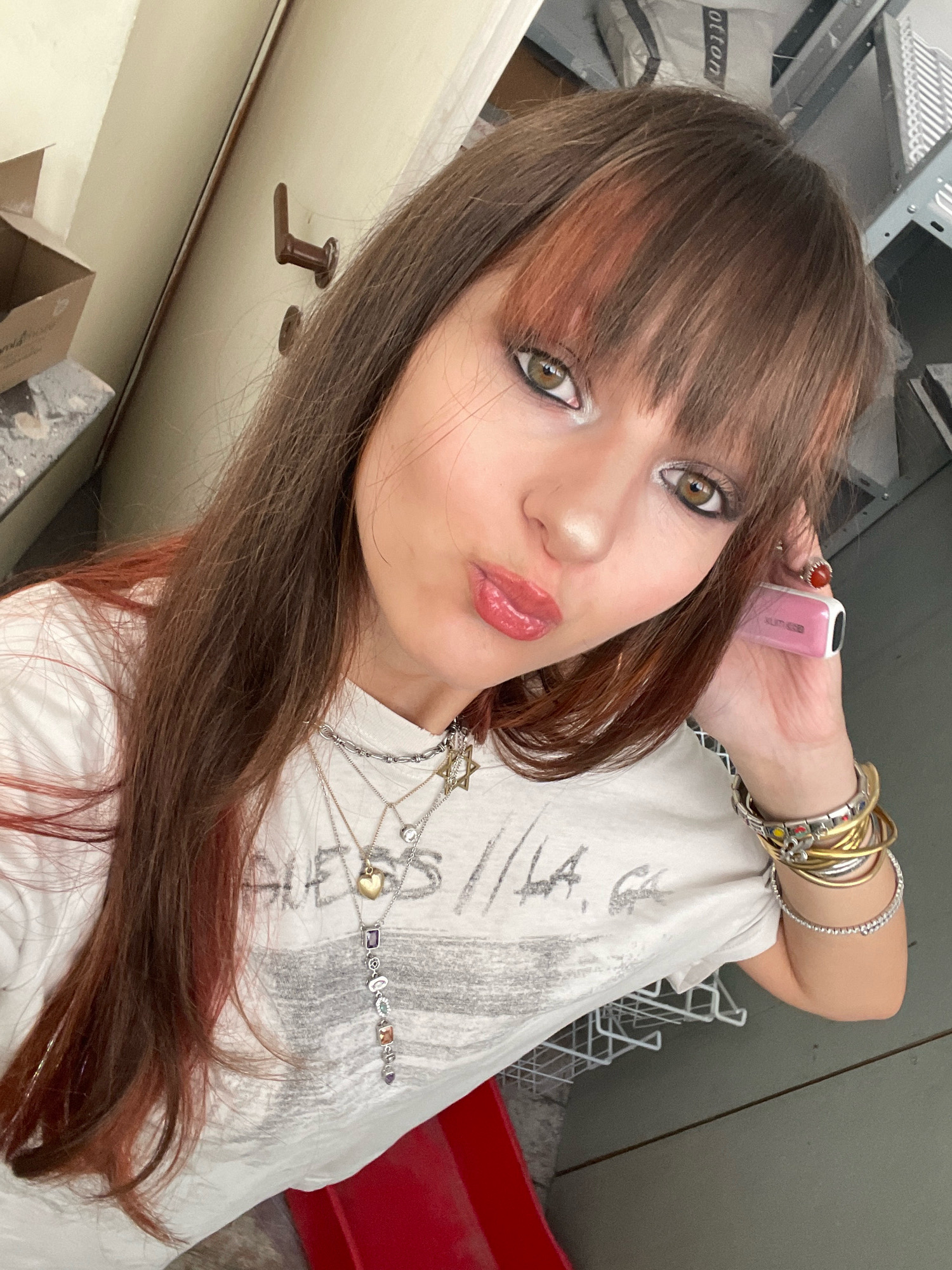 A young woman with brown hair, bangs, and light makeup poses for a selfie. She is wearing a white t-shirt with "GUESS L.A." printed on it and multiple necklaces. She also has several bracelets on her left wrist and a ring on her right index finger. A pink vape pen is held to her right ear. She is standing in front of a beige door with a brown handle. The background includes a cardboard box on a shelf, part of a metal shelving unit, and what appears to be a red object in the lower foreground. The scene is indoors, possibly a residential or storage area, and does not provide specific indicators of Raanana, Israel.
