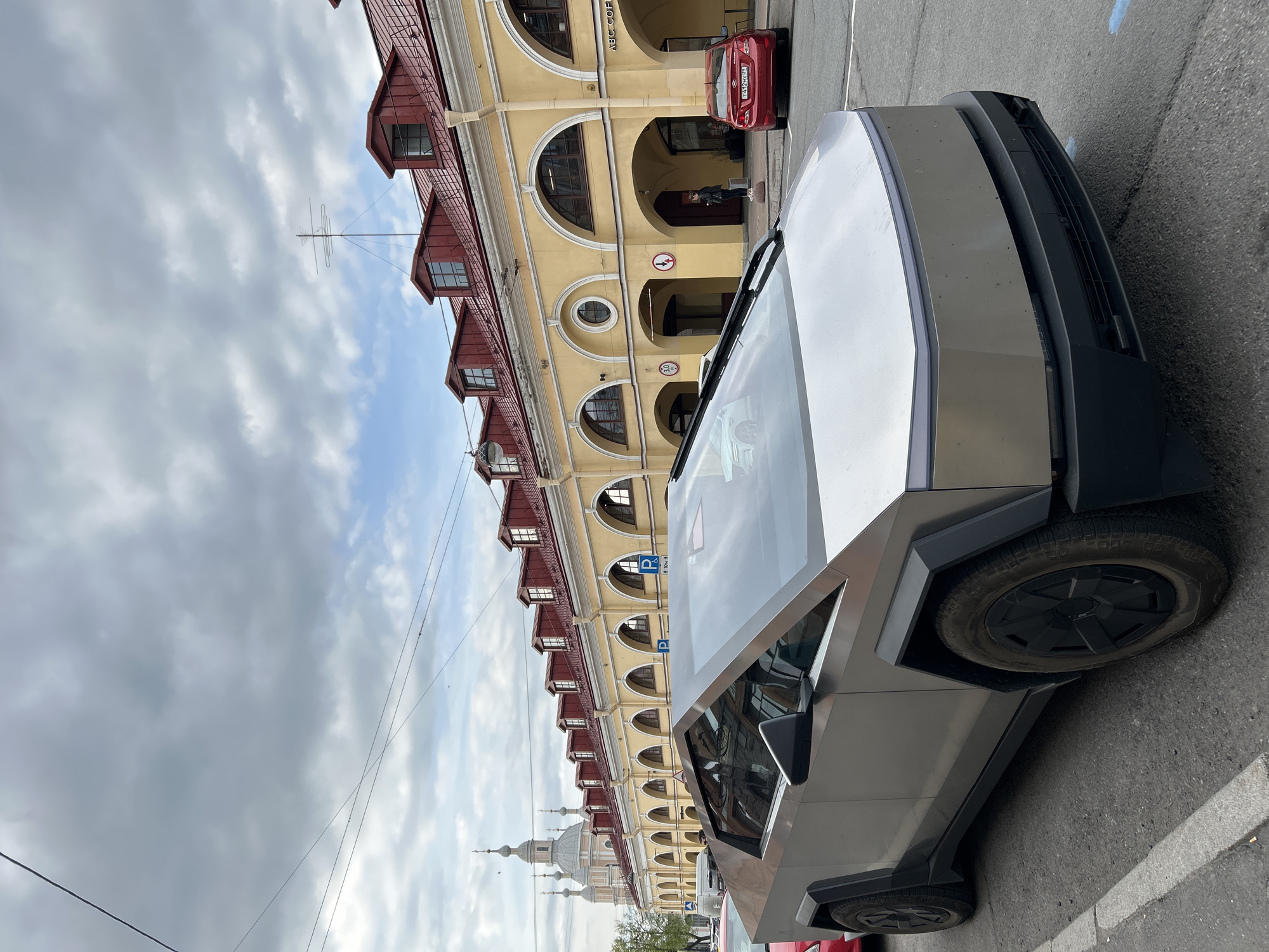 A Tesla Cybertruck is parked on a street in Saint Petersburg, Russia, in front of a yellow building with red roofs and arched windows. The building appears to be a historic structure. In the background, a church spire is visible. There are other vehicles parked nearby, including a red car. The sky is overcast with cloudy weather.