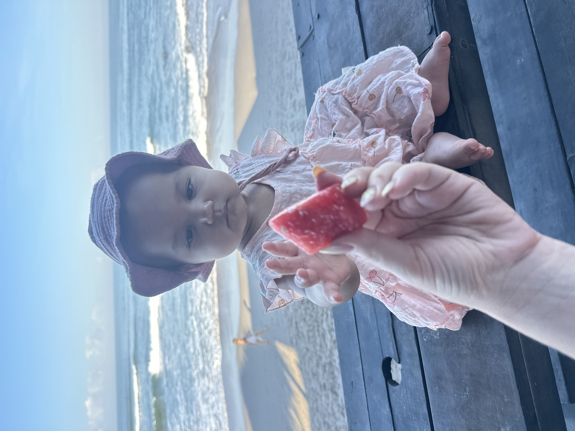 In North Kuta, Indonesia, a baby wearing a pink bucket hat and a frilly pink dress sits on a dark wooden table. The baby is reaching out a hand towards a piece of watermelon held by an unseen person. The background shows a beach with gentle waves and a clear blue sky.