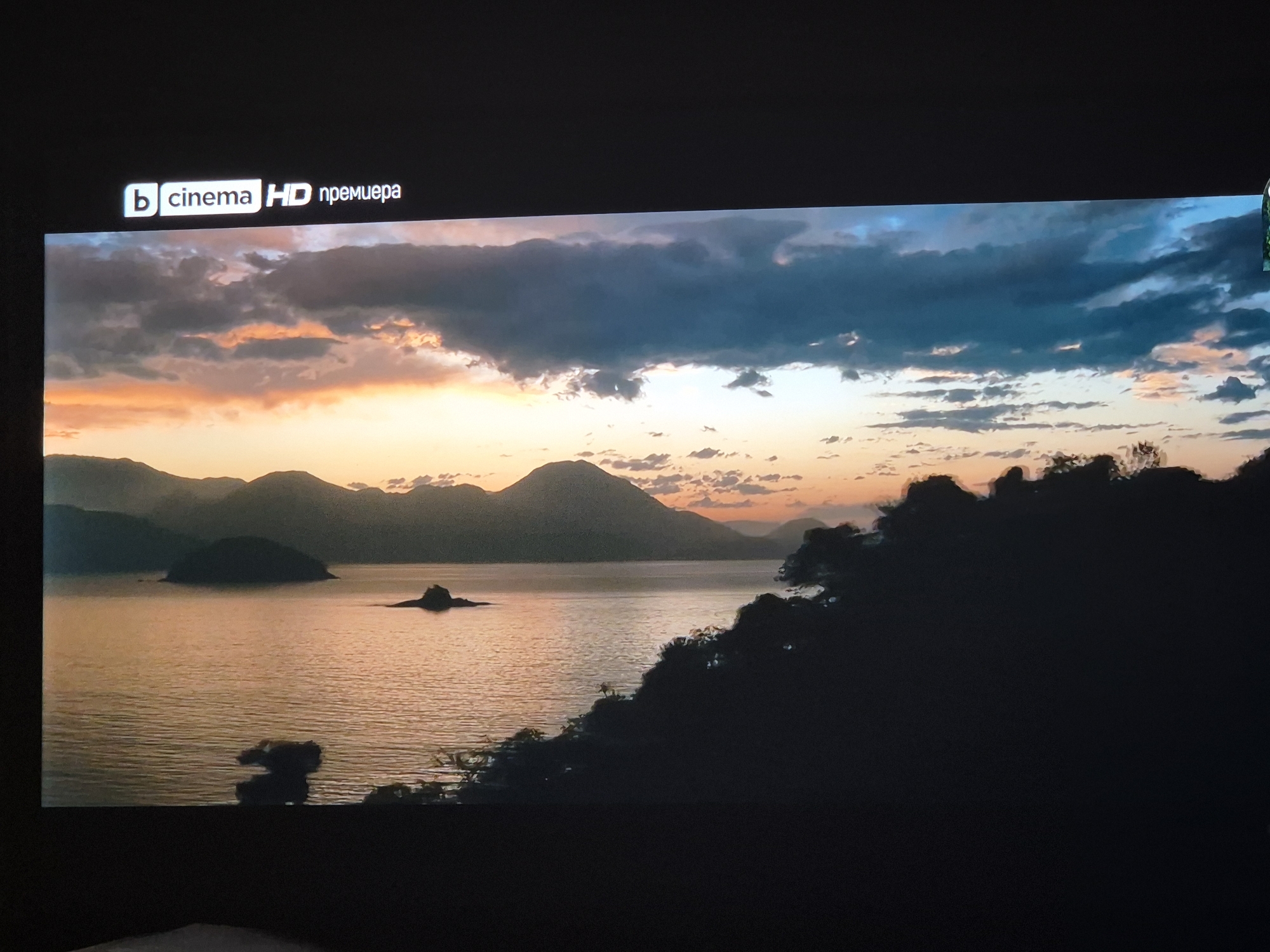 The image captures a beautiful cinematic landscape during what appears to be either sunset or sunrise. The scene features a calm body of water, likely a lake or a bay, reflecting the soft, warm light from the sky. In the mid-ground, several dark, rolling mountains or large hills are silhouetted against the vibrant horizon, their peaks gently fading into the atmospheric perspective.
The sky is a prominent feature, showcasing a dramatic blend of colors. On the left, the clouds and sky glow with hues of orange, pink, and soft yellow, indicating the setting or rising sun. Towards the right, the sky transitions into cooler tones of deep blue and grey, with scattered clouds casting soft shadows. A small, dark island or rock formation is visible in the middle of the water, and another smaller one is further to the right. The immediate foreground and the right side of the image are dominated by dark, silhouetted landmasses covered in dense foliage, adding depth and framing the serene vista.
In the top left corner of the screen, a digital overlay is visible. It features a logo with a stylized 'b' inside a rounded square, followed by the text "cinema HD". Next to this, in Cyrillic script, is the word "премиера", which translates to "premiere". This indicates that the image is a still from a broadcast or streaming service, possibly a movie or show being presented as a premiere on a cinema-themed high-definition channel. The overall mood is one of tranquil natural beauty, enhanced by the golden hour lighting.