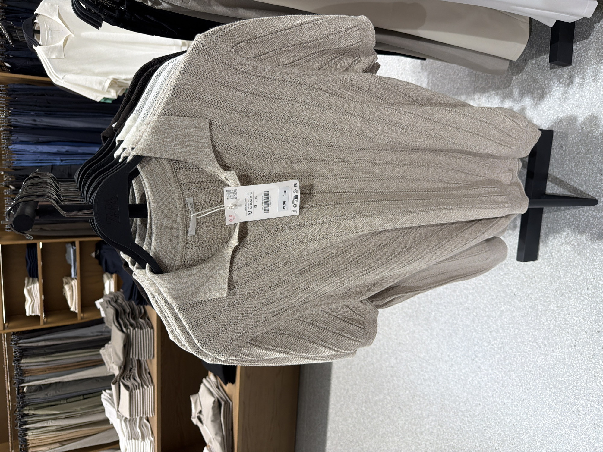 The image shows a close-up view of a beige, knitted, short-sleeved polo shirt hanging on a black hanger in a retail store. The shirt has a ribbed texture and a collar. A price tag with "39.90 CHF" is attached to the shirt. In the background, other clothing items are visible on shelves and racks, including folded pants and shirts, as well as other garments hanging on hangers. The store appears to have a modern and organized display.
