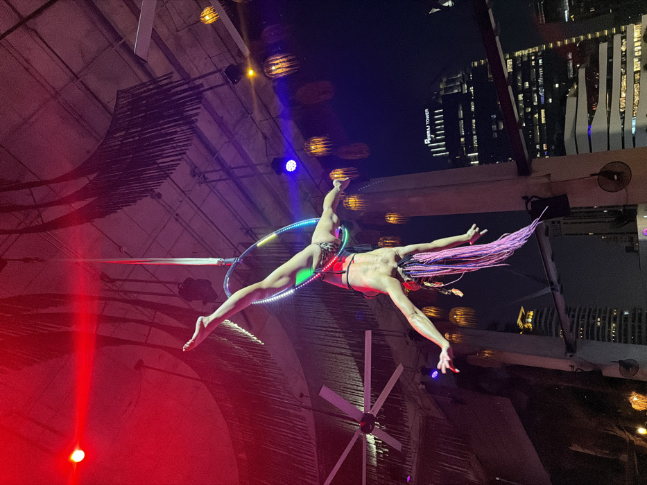 A female aerialist is performing an inverted pose on an LED-illuminated lyra, suspended by a white fabric strap from a high ceiling. She wears a leopard-print bikini bottom and a dark top, with long pink braided hair and red fingernails. Her left leg is extended vertically through the hoop, while her right leg is bent, and her arms are outstretched downwards. The overhead structure features exposed beams, dark decorative fringed fabric, and several spotlights casting red, blue, and purple light. Large white ceiling fans and multiple hanging globe-shaped decorative lights are also visible. In the background, against a dark night sky, numerous illuminated city buildings rise, with "OMNI TOWER" clearly legible on one prominent structure. The scene is an outdoor or semi-outdoor rooftop venue in Bangkok, Thailand.