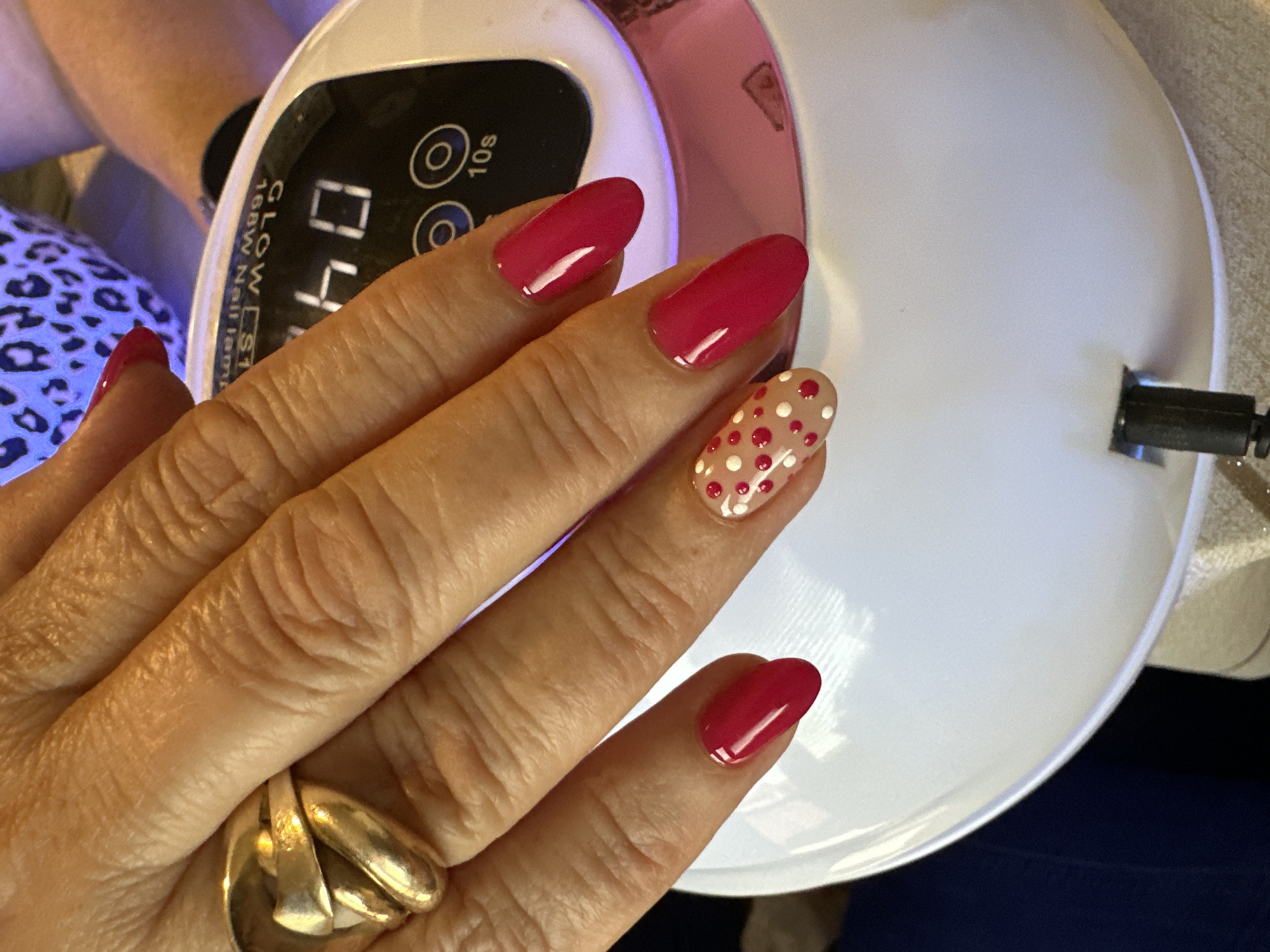 A close-up, high-angle shot features a person's left hand with a gold ring on the ring finger, positioned near a white and pink nail lamp. The fingernails are painted with a glossy, berry-red color, except for the middle fingernail, which has a light beige base with red and white polka dots. The nail lamp displays "04" on its digital screen, and buttons labeled "10s" are visible. The brand name, "GLOW ST" is printed on the device. The setting is indoors, and the scene is not located in Montana, Bulgaria, as the location is not visually discernible.