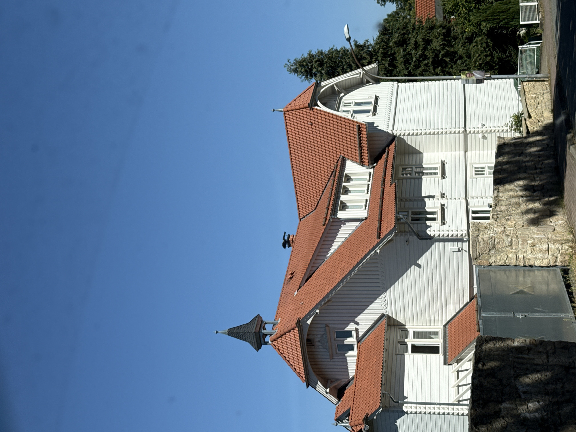 The image shows a white wooden house with a red tiled roof under a clear blue sky. The house has multiple gables and dormer windows. A small dark spire with a metal finial sits atop the highest roof peak. The exterior walls are clad in horizontal wooden planks with decorative trim. There are several windows visible, some with white frames. In the foreground, a stone wall and a metal gate are partially visible. To the right of the house, a tall evergreen tree stands, and a street lamp is positioned on the side of the road. A poster with a man's face is attached to the street lamp pole. The setting appears to be a residential area in Bad Sachsa, Germany.