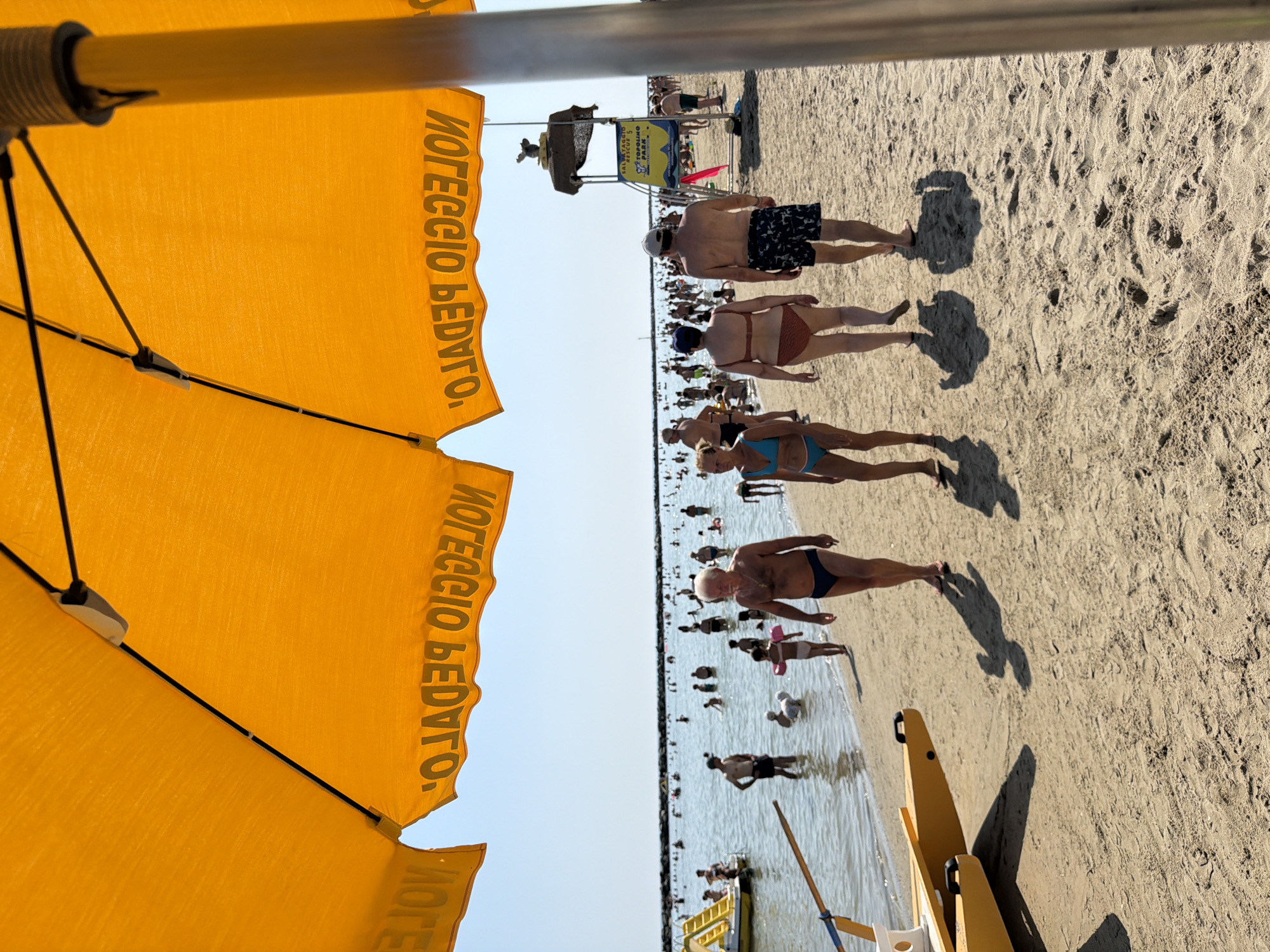An outdoor daytime shot shows a crowded beach in Gatteo, Italy, on a sunny day. The perspective is from beneath a large orange beach umbrella with the words "NOLEGGIO PEDAL" printed on its edge. In the foreground, a yellow and white rescue boat is partially visible on the sandy shore, with its shadow cast on the sand. Several people are walking on the beach, and a lifeguard tower stands further down the beach in the background, adorned with a sign that reads "SALVATAGGIO RESCUE 5 TOPOLINO PARK". The sea is calm and filled with numerous people swimming and standing in the shallow water. The sky is clear blue.