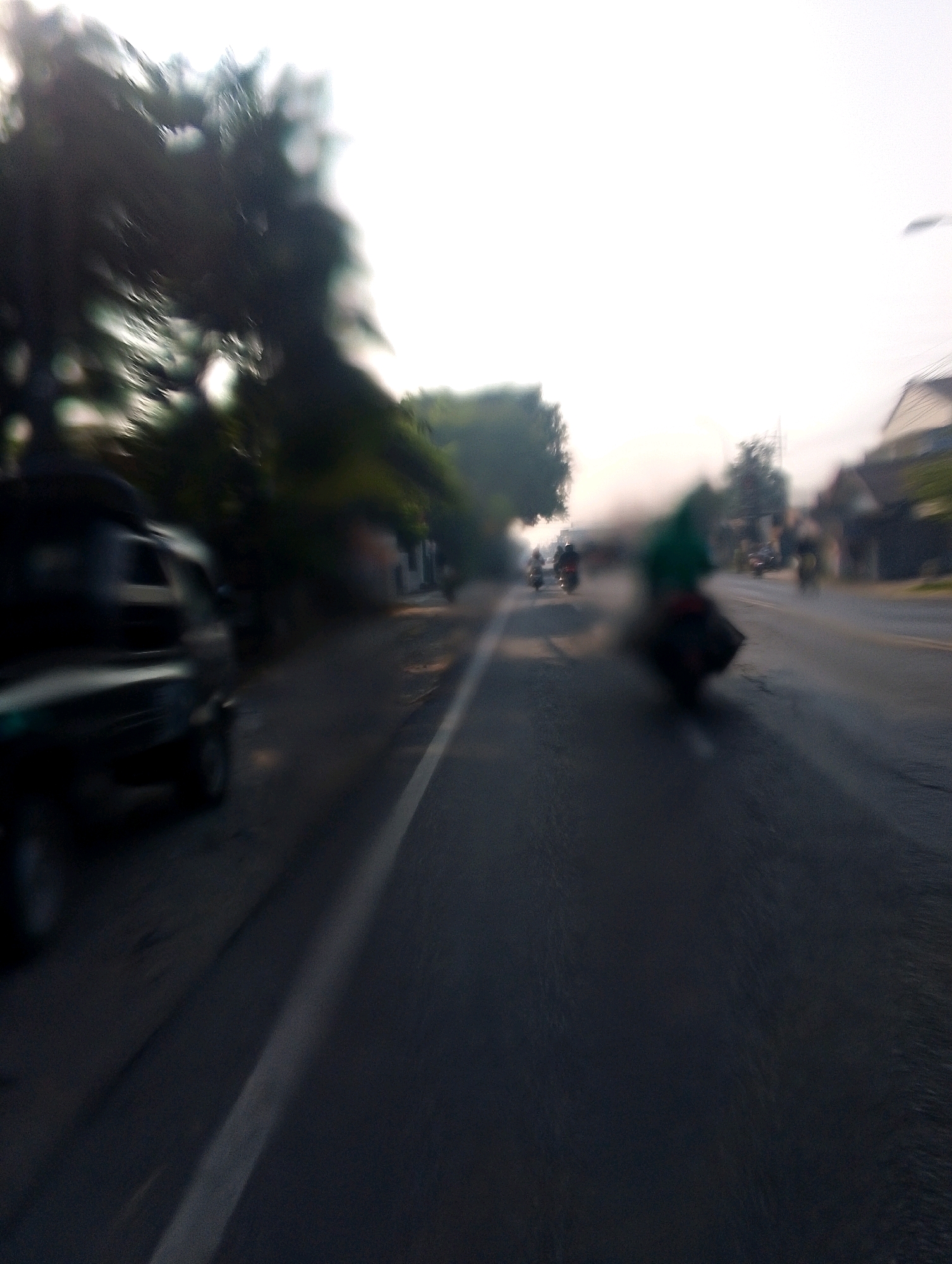 The image depicts a street scene with motion blur, suggesting a vehicle is in motion. On the left side of the road, a dark-colored vehicle, possibly a car or van, is partially visible. The road itself is asphalt with a white line marking the lane. Several motorcycles with riders are scattered along the road in the distance, also appearing blurred due to motion. Some trees and buildings are visible on the sides of the road, but their details are indistinct due to the blur. The sky is bright, suggesting daylight, possibly early morning or late afternoon due to the soft lighting. The overall impression is of a busy street with traffic in an urban or semi-urban environment.
