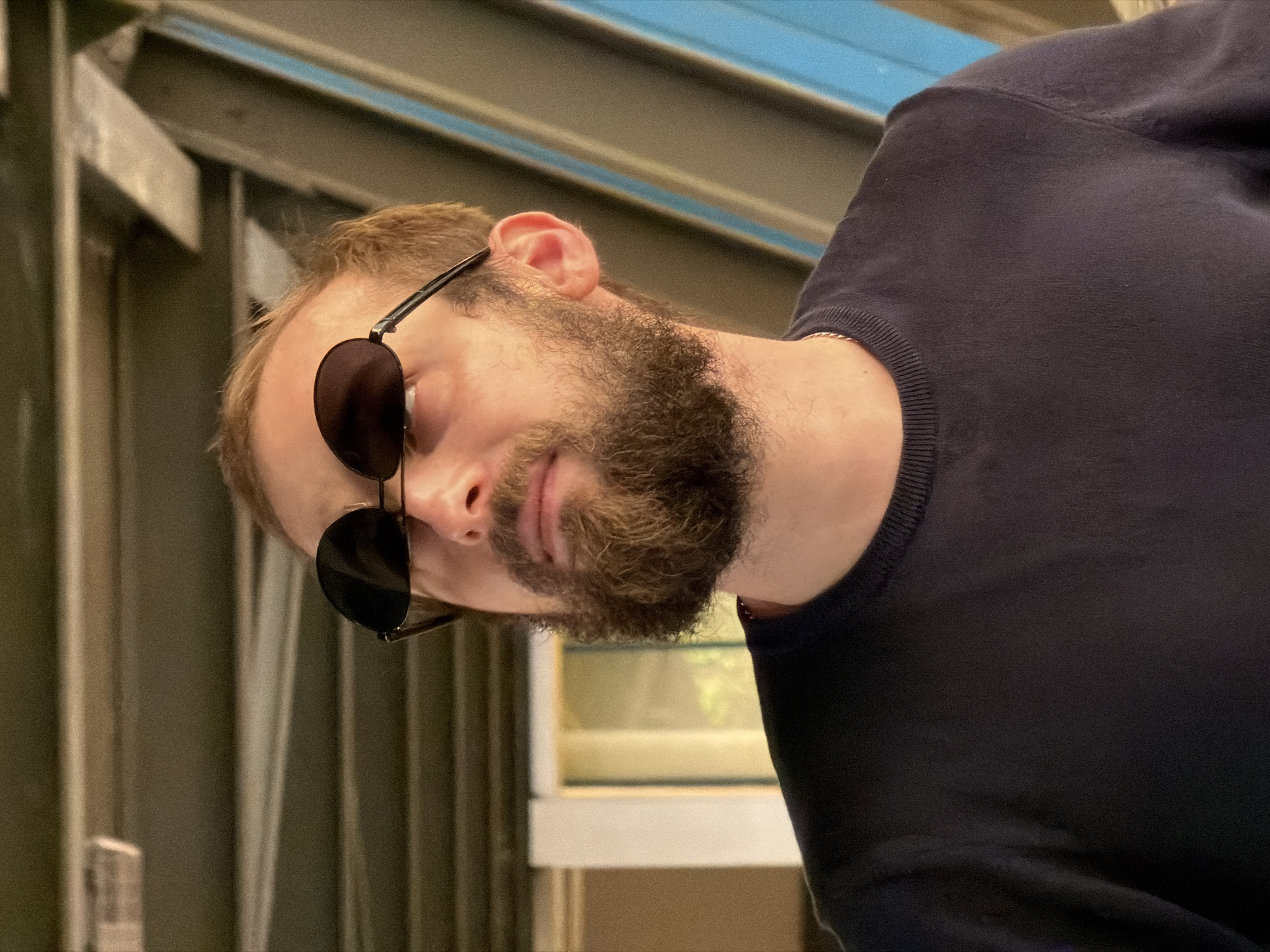 A man with a beard and sunglasses is wearing a dark blue sweater. He is tilting his head to the side. The background appears to be an urban environment, possibly in Moscow, Russia, based on the architectural elements. The scene includes a building facade.