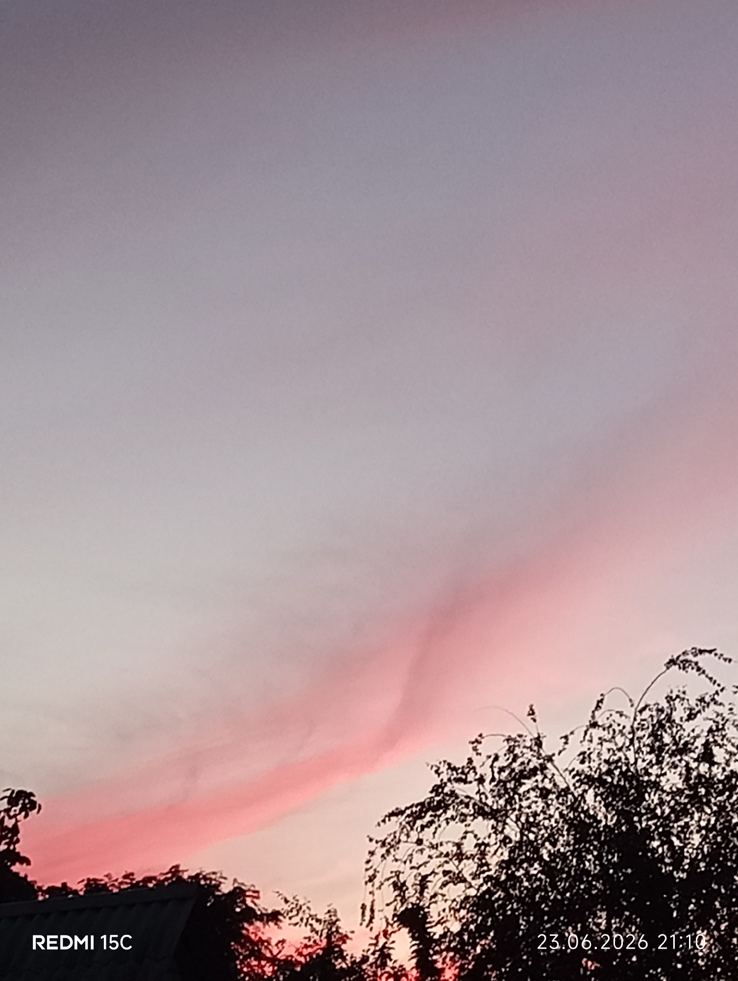 This is a medium shot of a sunset over Mykolaivka, Ukraine. The sky is a gradient of soft blues and purples, with a striking streak of bright pink cloud cutting across it. Silhouetted against the colorful sky are the tops of trees and the dark outline of a roof. The scene evokes a sense of peaceful twilight. The photograph was taken at 21:10 on June 23, 2026. Text on the image indicates the device used, "REDMI 15C".
