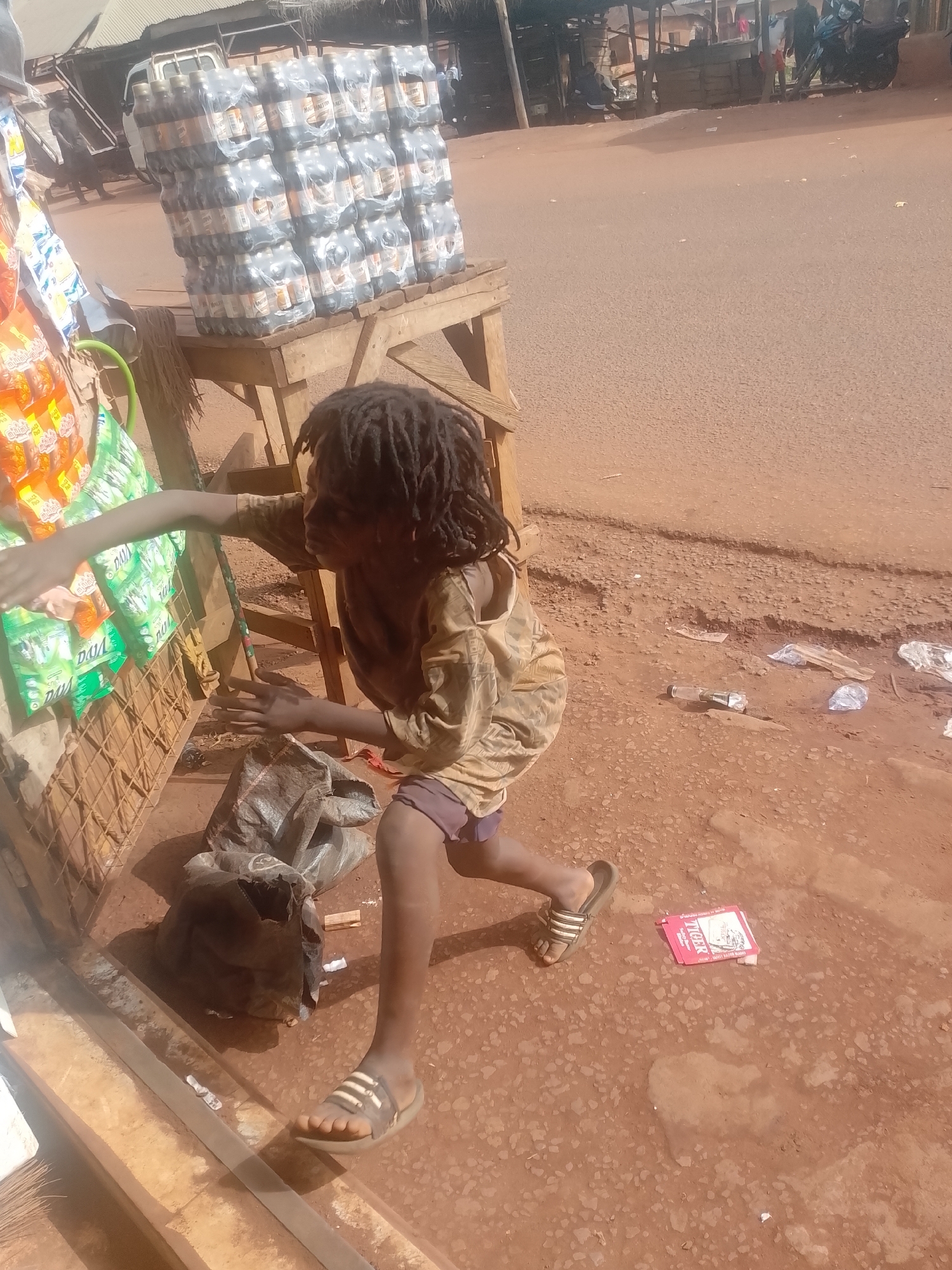 A young person with dreadlocks stands near a roadside vendor's stall in Ejule, Nigeria. The stall displays packaged goods, and a stack of bottled drinks is visible on a table behind the child. The ground is reddish-brown dirt, with scattered litter including a red packet. The person is wearing sandals and a light-colored shirt. In the background, the street scene includes other buildings and vehicles.