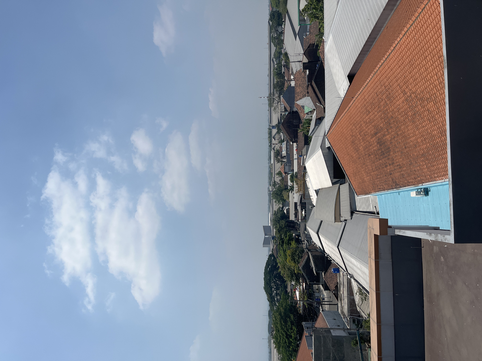 This is an aerial view of a residential and industrial area, likely in the city of Kedungdowo, Indonesia, on a clear, bright day. The foreground is dominated by the roofs of buildings. A prominent, reddish-orange tiled roof occupies the lower right portion of the frame, showing some signs of wear and discoloration. To its left is a section of a dark gray building, with a lighter gray horizontal band and a turquoise-colored metal roof with a white air conditioning unit attached. Behind these, a long, low-profile gray metal roof stretches across the mid-ground, interrupted by other buildings with a mix of gray metal and terracotta tiled roofs.
The mid-ground reveals a densely packed neighborhood with houses featuring various roof styles, some with peaked designs and others with flatter profiles. Streets are visible winding through the neighborhood.
In the distance, a line of trees, including a large, mature tree with a wide canopy, separates the residential area from industrial or commercial structures. Further in the background, a large, white, modern building with a distinctive angular design stands out. Beyond this, a long, low-slung industrial building or complex extends across the horizon, with some taller structures, possibly smokestacks or antennas, visible. The overall landscape appears relatively flat, with a hazy horizon suggesting a significant distance. The sky is a pale blue with scattered white, fluffy clouds. There are no people visible in the image.