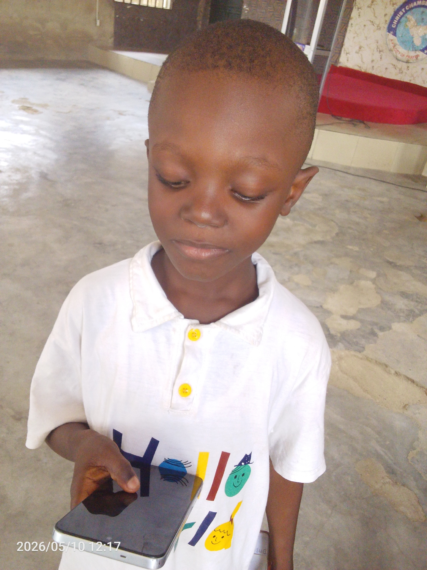 A young Black child stands indoors, holding a smartphone in their left hand. The phone screen is black and reflective. The child is wearing a white polo shirt with yellow buttons and colorful cartoon graphics on the front. The location appears to be inside a building, possibly a church, given the presence of a sign with the words "CHRIST CHAMBER" in the upper right corner and a red cushion near the sign. The background is neutral-colored, with a concrete floor. There is no indication that this image was taken in the city of Lokoja, Nigeria.