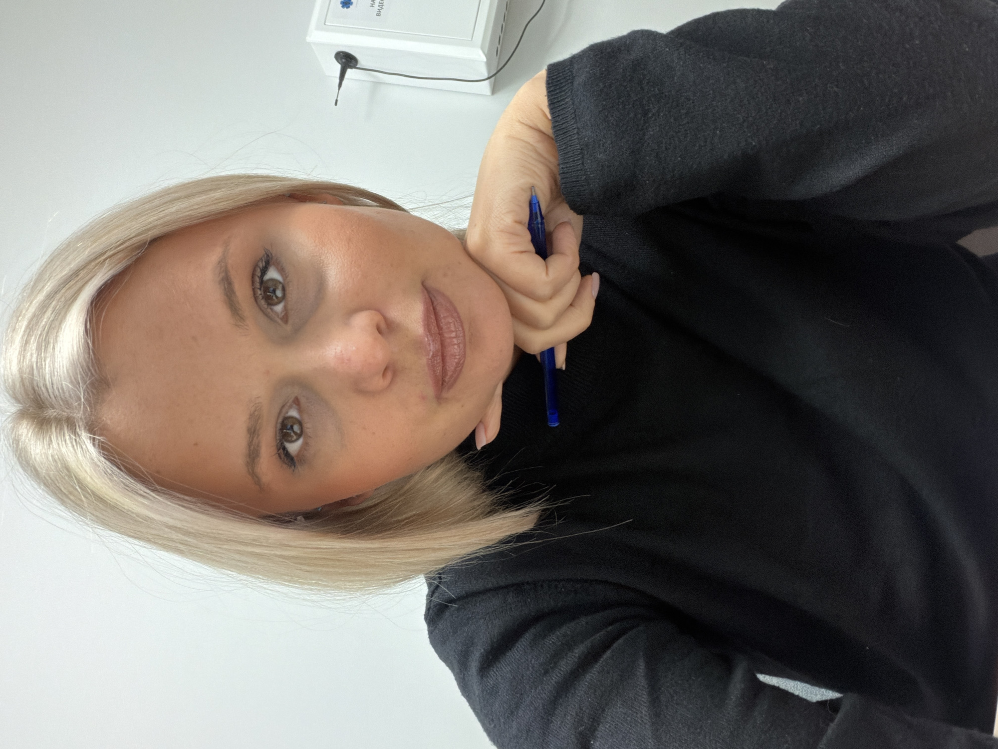 A woman with blonde hair is holding a blue pen in her right hand, which is resting on her neck. She has light skin, brown eyes, and is wearing a black turtleneck sweater. The background is a white wall with an electronic device attached to it. There is no evidence to indicate the location is Sarapul, Russia.