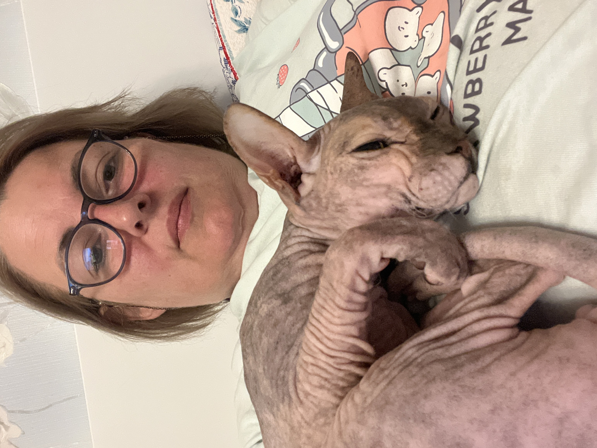 A person with blonde hair and glasses is lying down, holding a Sphynx cat. The cat is curled up on the person's chest, appearing relaxed. The person is wearing a light-colored t-shirt with a cartoon strawberry and bunny graphic. The background is a white wall with a subtle textured pattern. The scene appears to be indoors. The image was taken in Korolyov, Russia.
