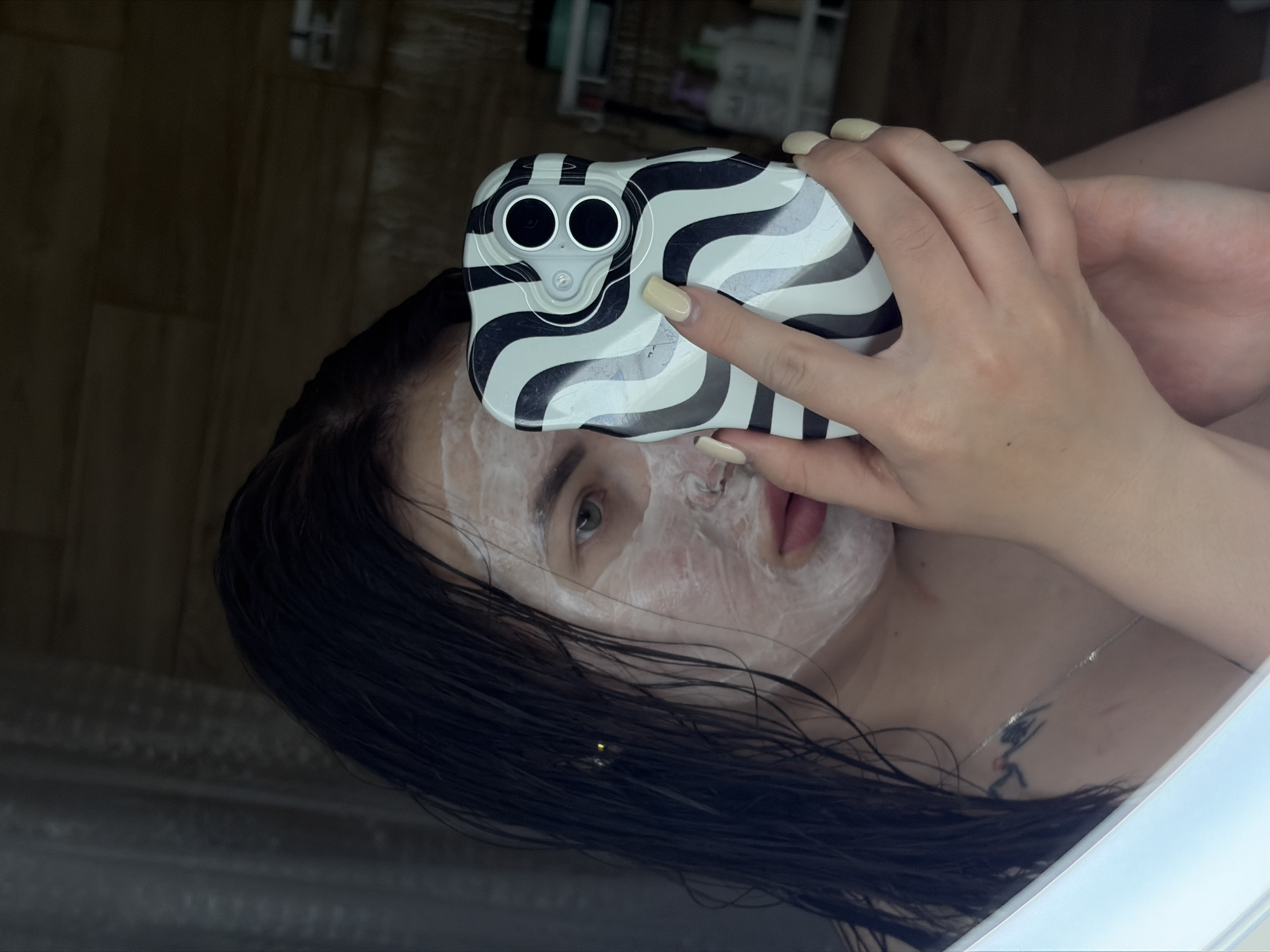 A person with a white facial mask is holding a phone in front of their face. The phone has a black and white patterned case and two visible camera lenses. The person has wet, dark hair and fair skin. A tattoo is visible on their upper chest. The background appears to be a bathroom, with a dark wood-like wall partially visible. The exact location in Krasnodar, Russia, cannot be determined from the image.