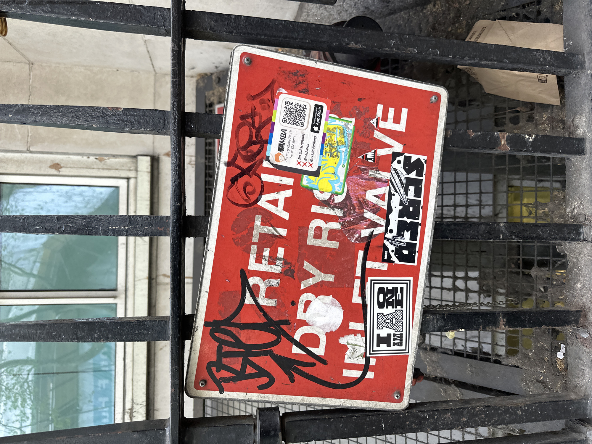 The image shows a weathered red sign attached to a black metal grate. The sign is heavily marked with graffiti and stickers. The primary words visible on the sign are "RETAIL", "DRY RIS", and "VALVE". There are stickers on the sign, with one displaying a QR code and text promoting an app called "AMBA". Another sticker reads "SECRET". A third sticker is white and black and includes the text "I AM ONE HETAY". The sign is surrounded by the bars of a black metal grate, and in the background, a beige building with a window reflecting the outside is visible. Below the grate, a brown paper bag is visible. The location appears to be an urban environment.