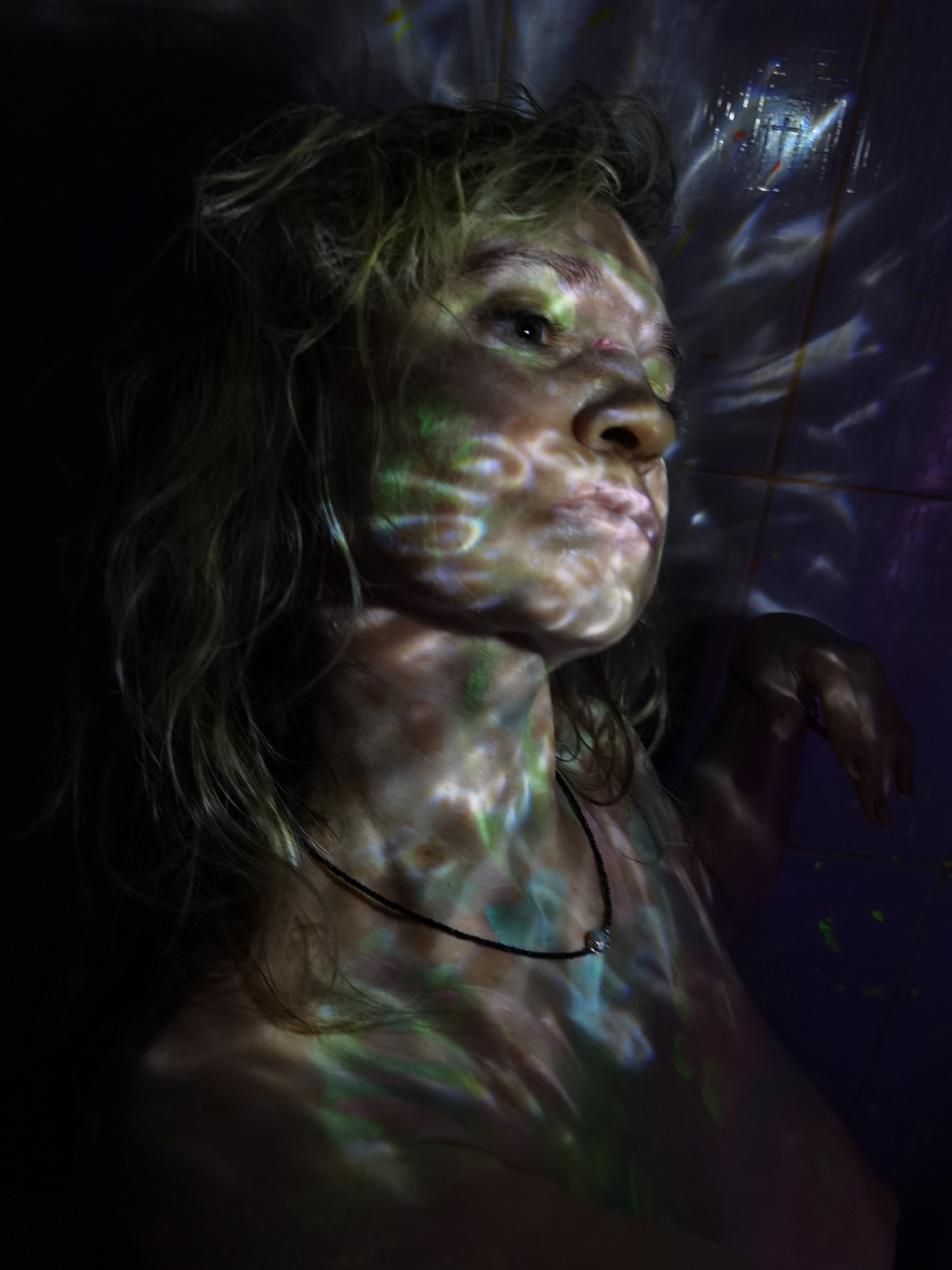 A close-up portrait features a person, likely a woman, illuminated by colorful projected light patterns against a dark background. Her head, neck, and upper torso are visible, with her gaze directed towards the upper right, giving her a contemplative or serene expression.
The person has light-colored, wavy hair that frames her face and shoulders. The dominant visual element is the abstract, swirling light patterns, primarily in shades of green, blue, and white, which are cast onto her face, neck, chest, and hair. These patterns create dynamic highlights and shadows, accentuating her features and giving her an ethereal appearance, reminiscent of light reflecting off moving water.
She appears bare-shouldered, wearing a thin black cord necklace adorned with a small, reflective bead or pendant around her neck. Her right hand and forearm are visible on the right side of the frame, resting against a dark surface that also catches some of the projected light. The background is mostly dark, with a dark purple or blue wall visible on the right, showing similar, diffuse light patterns and faint tile lines. The overall impression is one of artistic lighting and a dreamlike, intimate atmosphere in a dark indoor setting. There is no visible text.