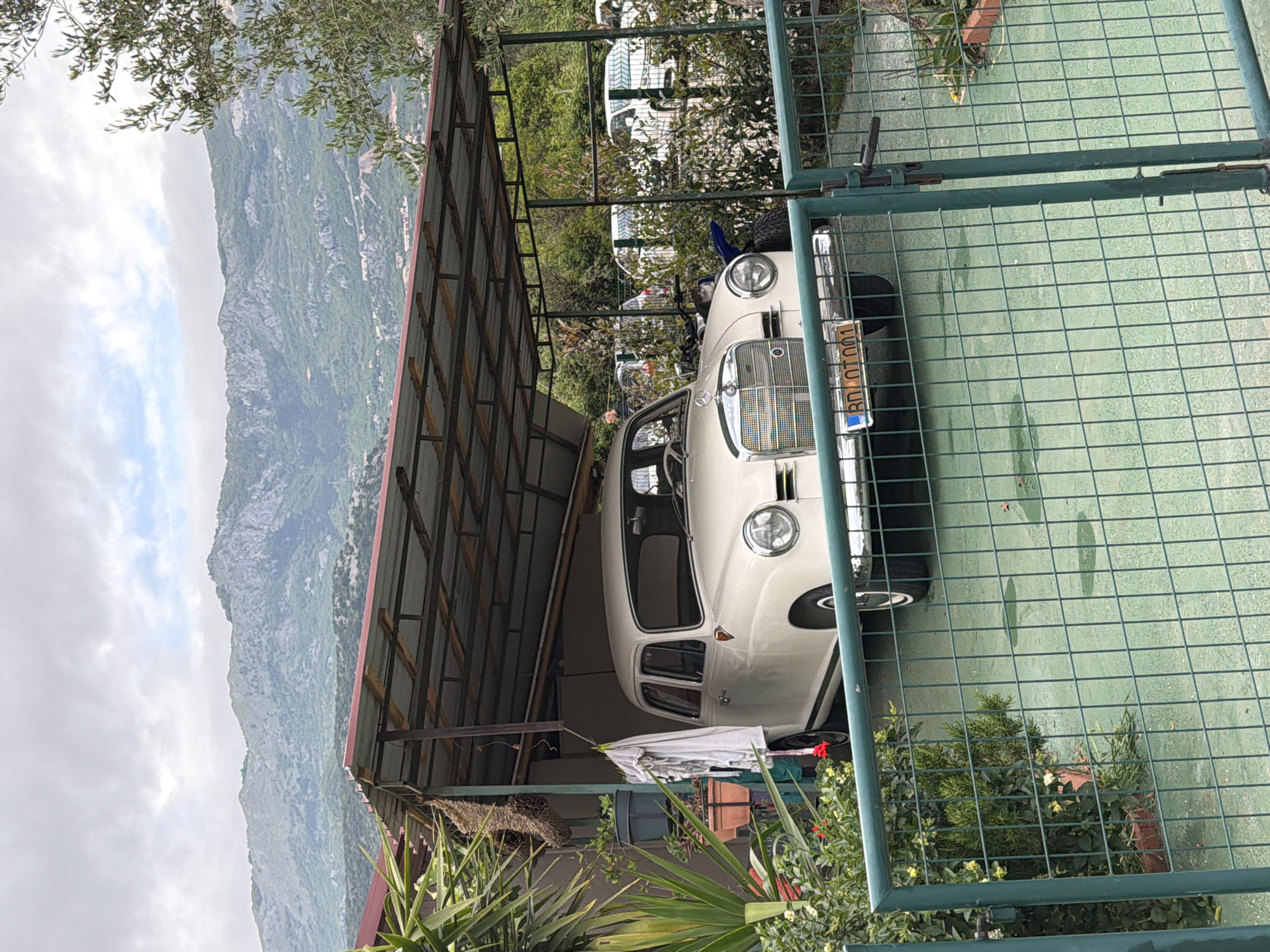 The image shows a light-colored vintage Mercedes-Benz car parked partially under a metal roof structure. The car has a license plate that reads "BD/AT/001." The car is partially blocked by a green wire fence in the foreground. Behind the car, there is lush vegetation. Beyond the car, metal roof, and vegetation is a view of a mountain range partially obscured by clouds. The location is suggested to be Budva, Montenegro. No people are visible.