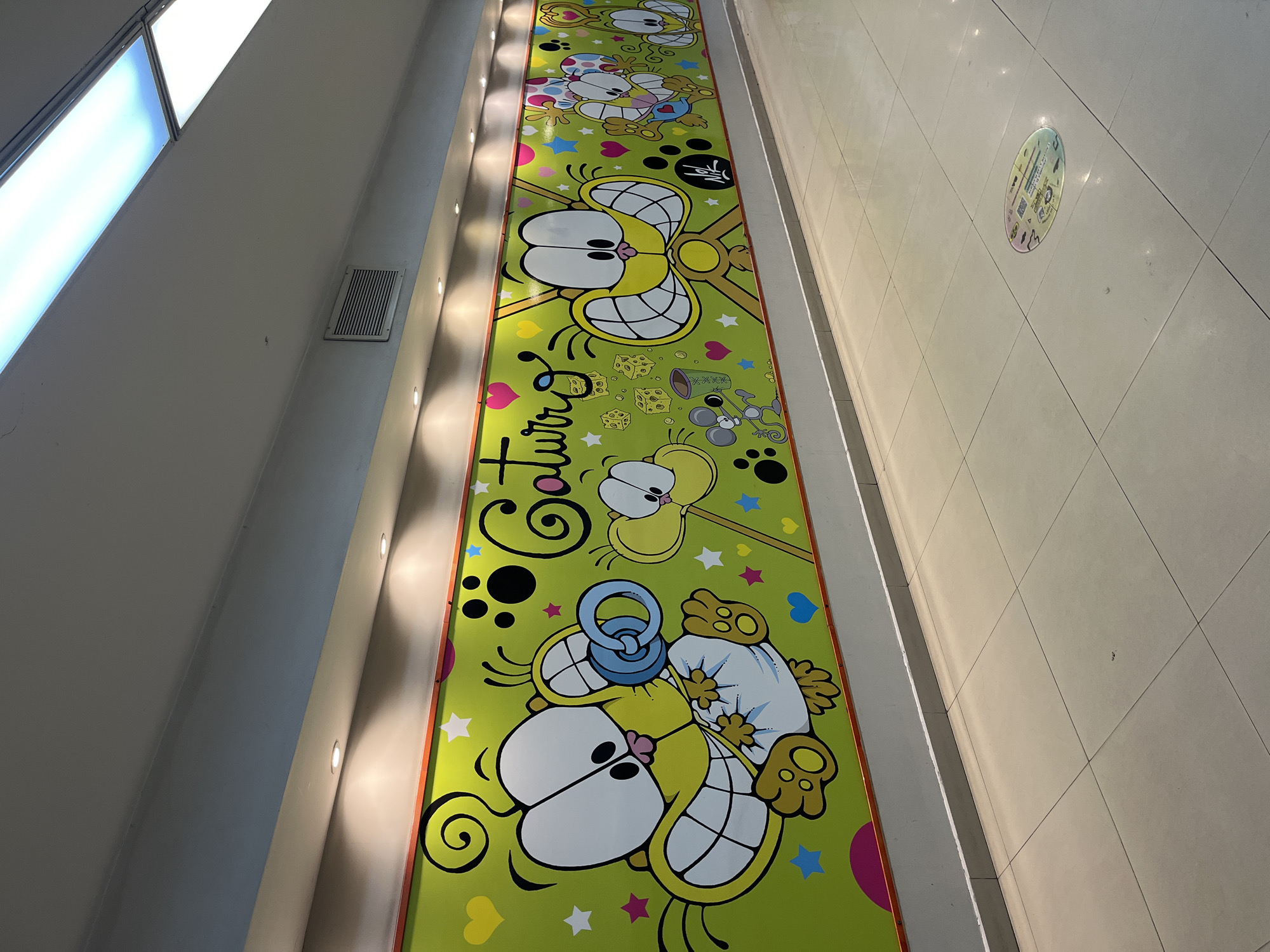 This media file captures a long, brightly lit indoor corridor in Buenos Aires, Argentina, adorned with a vibrant floor-to-ceiling mural. The dominant feature is a lime-green mural spanning the length of the corridor, depicting several stylized yellow cat-like characters, strongly reminiscent of the Argentine comic strip character Gaturro.
The mural showcases these playful cats in various forms: one with multiple eyes, another as a baby wearing a diaper and a blue pacifier. A grey mouse character, surrounded by scattered cheese cubes and a cup, also appears. The background is embellished with colorful stars, hearts, and swirling black patterns.
The corridor itself features modern architecture with clean lines. Recessed spotlights illuminate the mural from a bulkhead above, while large rectangular fluorescent lights provide overhead lighting on the opposing wall. An air vent is visible on the wall. The floor is covered with light-colored square tiles, and further down the corridor, a circular, iridescent sticker with small icons and illegible text is placed on the ground.
Prominently featured on the mural is the stylized word "Gaturro" in a bubbly font, appearing multiple times. Some illegible characters in an East Asian script are also present above one of the cat figures.
The overall scene conveys a cheerful and whimsical atmosphere, likely designed to entertain or guide visitors, possibly in a mall or public building. The image is taken from an elevated, angled perspective, looking down the length of the decorative corridor.
