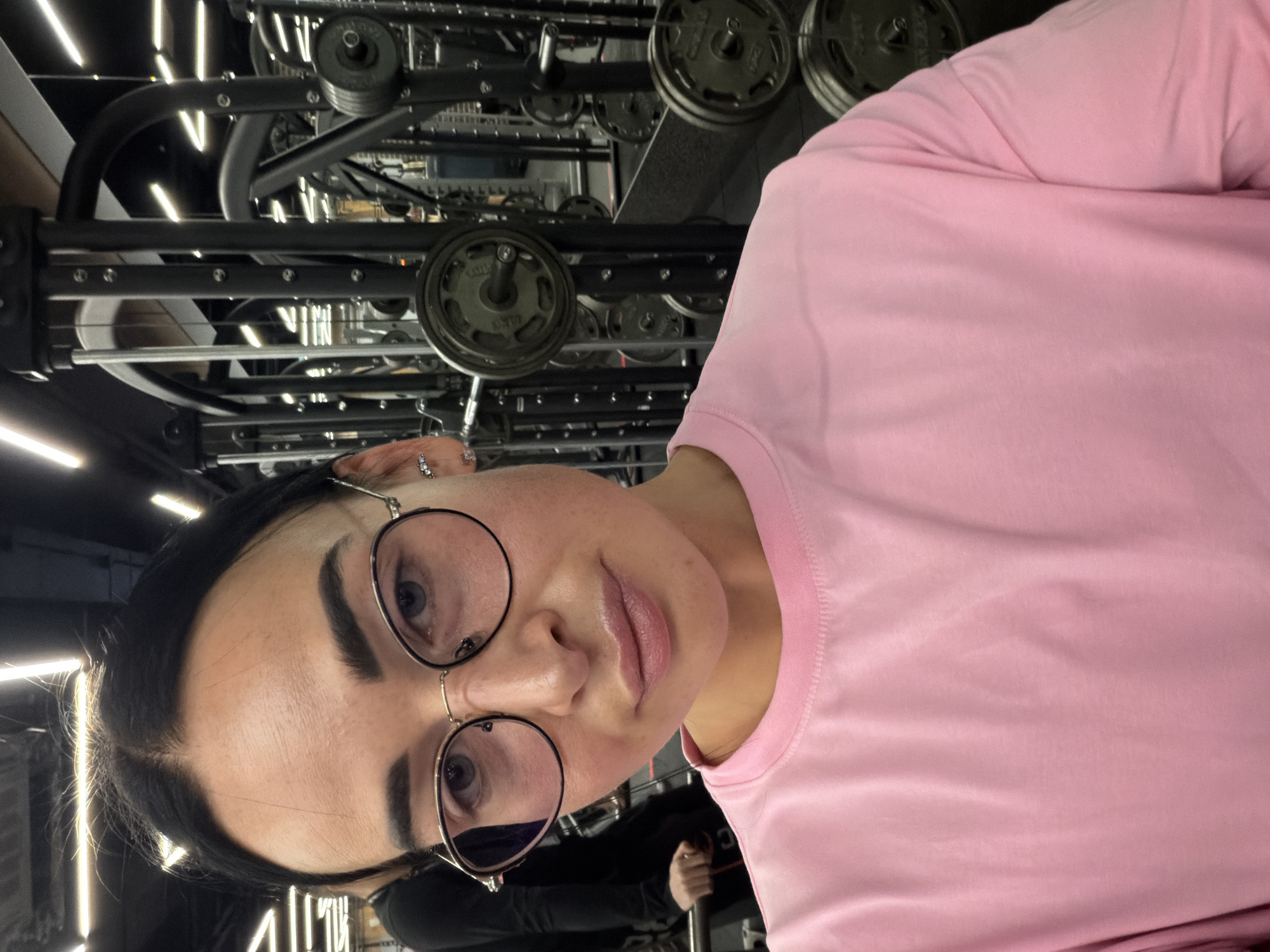 A woman wearing a pink shirt and glasses is positioned in front of a weight lifting machine. The surroundings appear to be a gym setting. The location cannot be determined as Saint Petersburg, Russia, from this image alone.