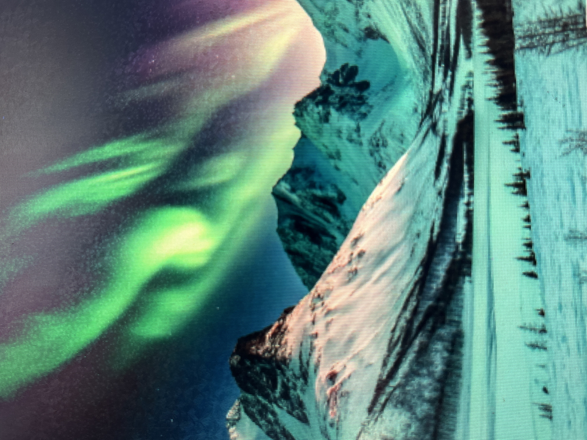 The image displays a vibrant aurora borealis illuminating a snowy mountainous landscape. The sky is a deep blue, transitioning to purple and yellow hues where the aurora is most intense, with prominent green streaks of light dancing across the upper portion of the frame. Below the celestial display, snow-capped mountains dominate the midground, their peaks sharply defined against the twilight sky. The slopes are dusted with snow, revealing dark, rocky outcrops and sparse vegetation. In the foreground, a vast, snow-covered plain stretches out, dotted with a line of dark evergreen trees. The overall scene depicts a cold, serene, and natural environment. The city of Sejahtera, Indonesia is not depicted in this image, as it is a tropical country and the depicted landscape and phenomenon are not characteristic of its geography or climate.