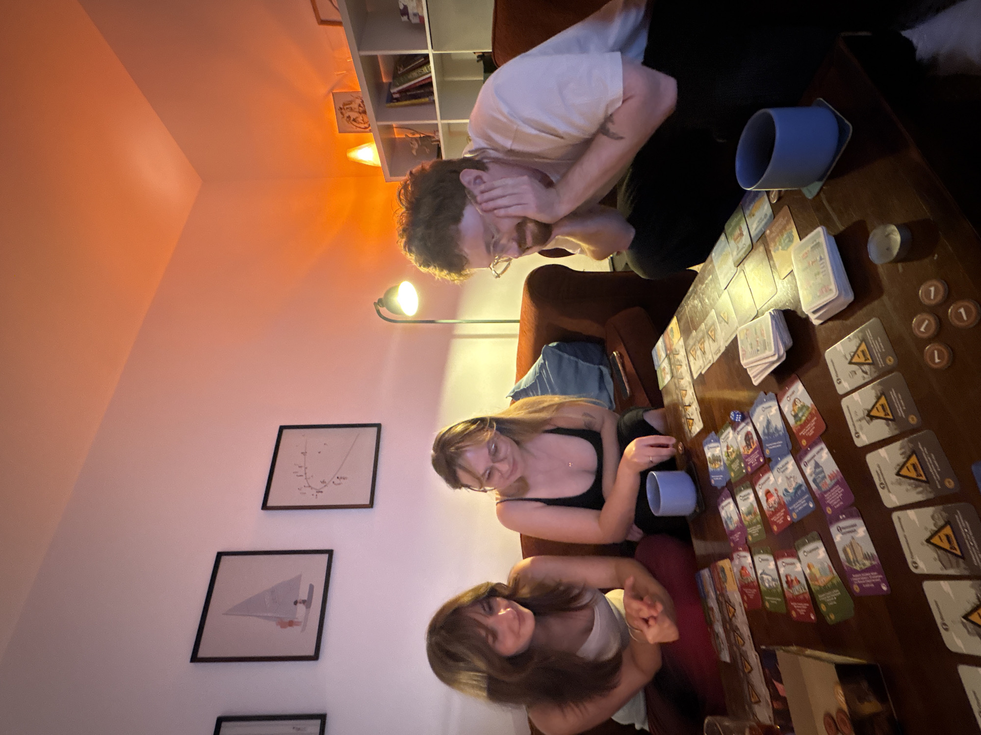 The image shows three people sitting around a table playing a board game. The person on the top left is a male with glasses, wearing a white t-shirt and black pants. He has his hands on his face, suggesting shock. Below him and to the right is a female wearing a black tank top. Below her is another female, wearing a white tank top and maroon pants. There are framed pictures on the wall behind them, and a bookshelf in the upper left corner of the image. The table is covered with cards, tokens, a die, and two blue mugs. The game appears to be Pandemic. The location is Ramat Gan, Israel.