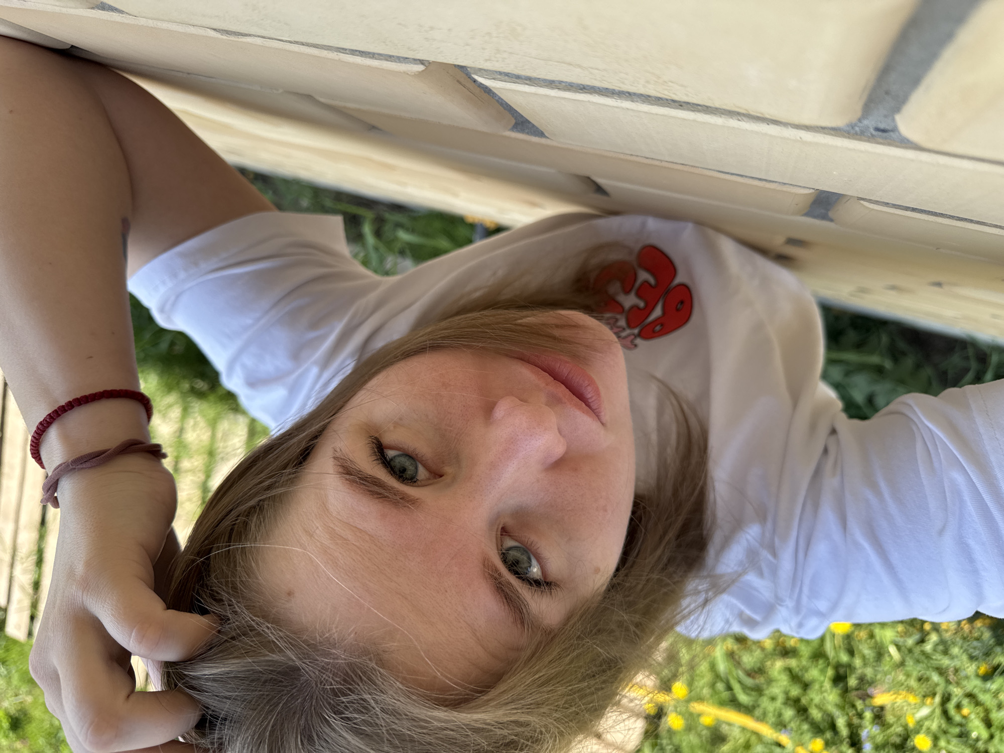 A young woman lies on her side on a grass-covered surface in what appears to be an outdoor setting. She is wearing a white T-shirt with a red design on the upper right. Her left hand is near her head, seemingly touching her hair, while her right hand is mostly out of view. She has fair skin, light brown hair, and green eyes, and she is looking directly at the camera. Behind her, a brick structure or wall is visible. Based on the location given, the image appears to be taken in the city of Ферма, Russia, but it's not possible to determine the specific place in the city.