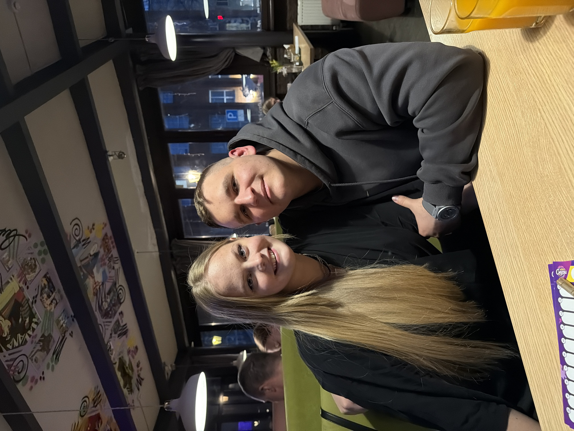 The image displays two young individuals, a man and a woman, seated at a light wooden table in what appears to be a cafe or restaurant during the evening. Both are looking towards the camera and smiling, conveying a cheerful mood.
The man, positioned on the left, has short dark hair and is wearing a dark grey hooded sweatshirt. A grey watch with a round face is visible on his left wrist. The woman, on the right, has long blonde hair flowing over her shoulders and is wearing a black top with a delicate necklace.
On the table in front of them are two glasses containing an orange beverage, likely juice. To the right of the table, a purple and white sheet of paper, possibly a menu or order form, displays the Russian word "СОУС" (Sauce) along with numbers and blank spaces.
The background features large windows revealing a dark outdoor scene with blurred city lights and reflections, including a faint blue 'P' parking sign. The interior is lit by modern pendant lamps with white shades. To the left, a wall section is adorned with colorful, abstract artwork. Green upholstered seating is visible beneath the woman. The ceiling has exposed dark beams and white panels. The scene suggests a relaxed social outing in Izhevsk, Russia.