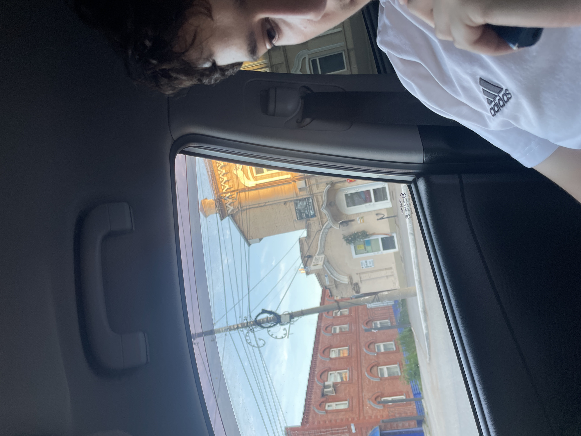 An individual wearing a white Adidas t-shirt is seated inside a vehicle, looking out the window. Through the window, a street scene in Votkinsk, Russia, is visible. The street is lined with old brick buildings and a building with a light-colored facade. A utility pole with numerous wires stands in front of the buildings. The sky is visible and appears clear. The time of day appears to be late afternoon or early evening due to the lighting. The image is taken from a slightly elevated perspective, possibly from the front passenger seat of the vehicle.