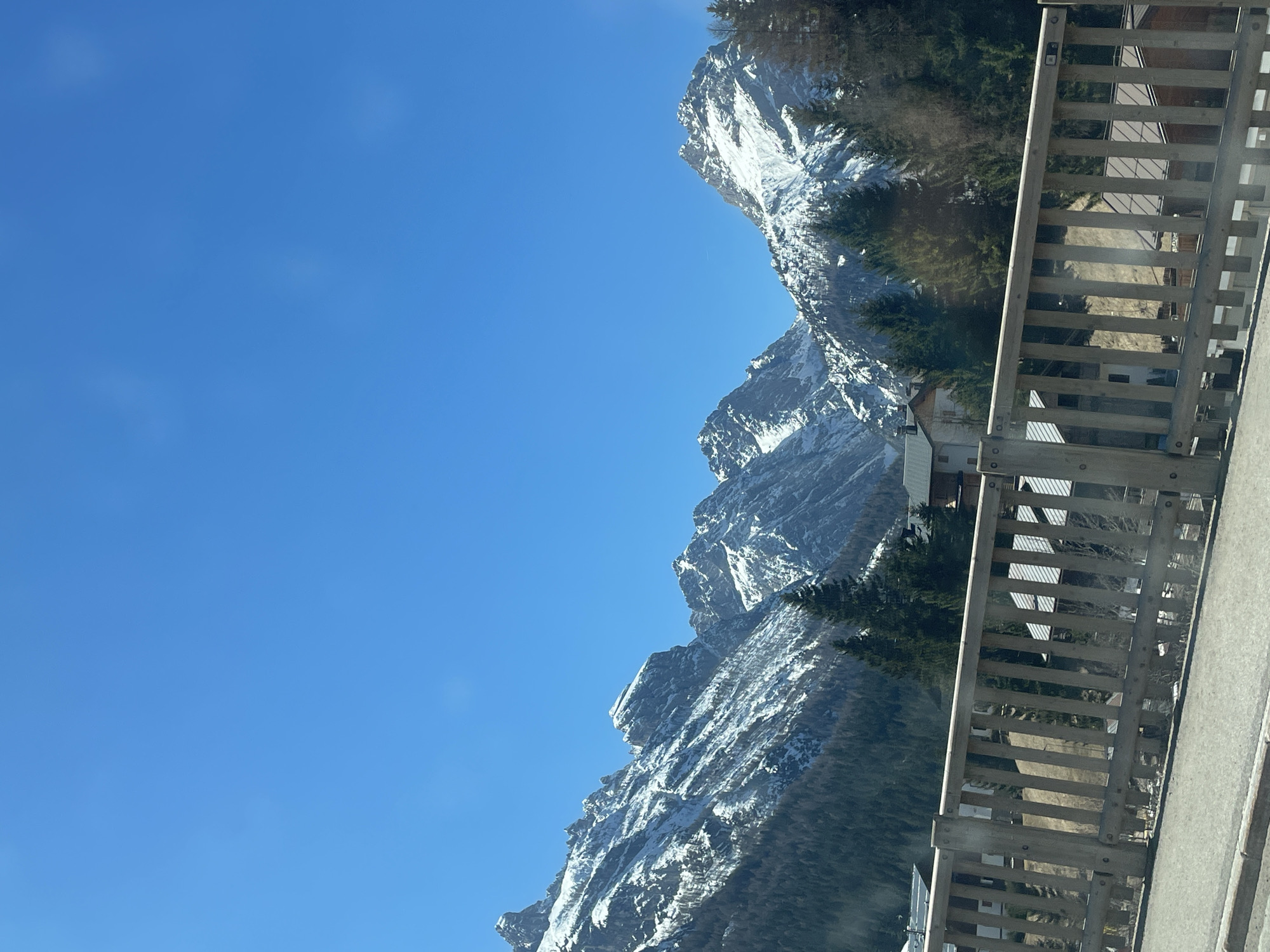 The image depicts a mountain range in Sappada, Italy. The mountains are covered in snow and surrounded by green trees. A wooden fence is visible in the foreground, obscuring part of the mountain range. The sky is blue and clear. Some rooftops of buildings are visible between the fence and the mountains.