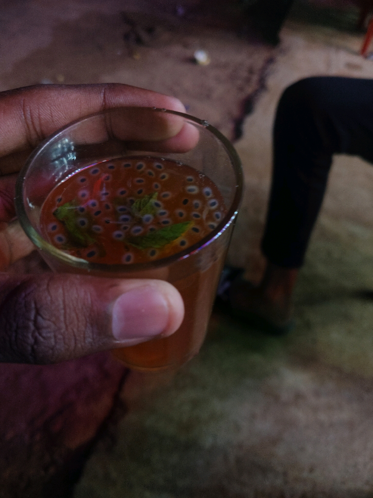 A hand holds a clear glass filled with a reddish-brown liquid containing basil seeds and mint leaves. The glass is partially submerged in shadow. In the background, a person in dark clothing is seated on the ground. The environment appears to be outdoors with a dusty, uneven surface.