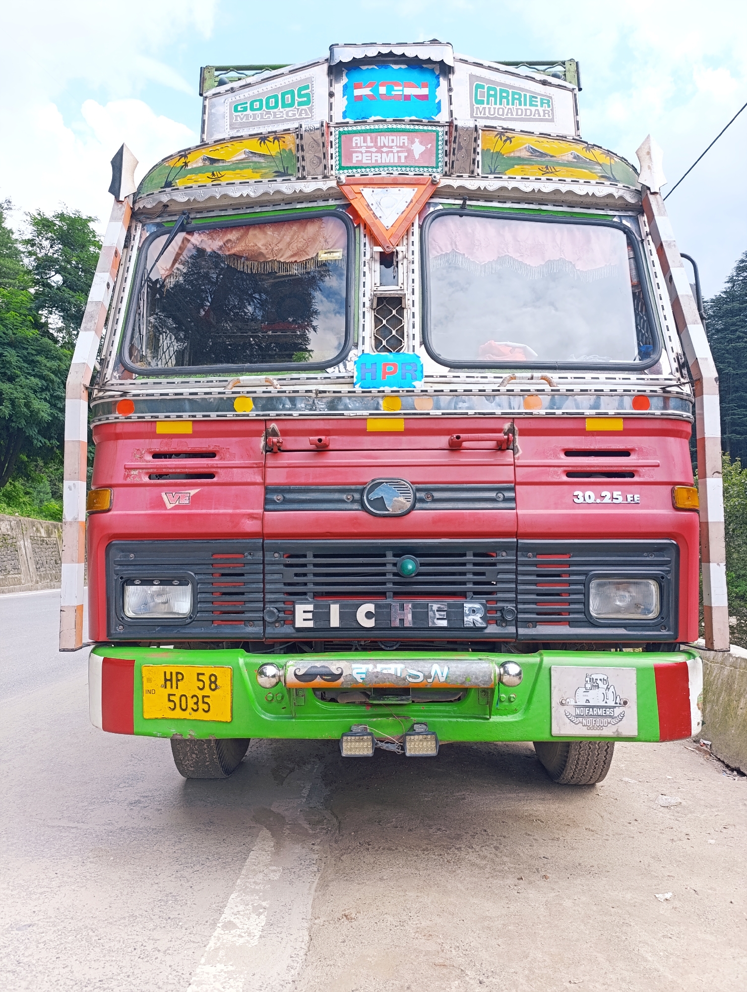 A front-facing, eye-level, medium shot captures a vibrantly decorated red and green truck parked on an asphalt road in Vashisht, India. The truck is heavily adorned with colorful decals and signage. At the top, a sign reads "GOODS MILEGA" and another says "ALL INDIA PERMIT" above a map of India. To the right, "CARRIER MUQADDAR" is displayed. The truck's front features a prominent chrome grille with "EICHER" inscribed. Below the grille, a bright yellow license plate reads "HP 58 5035". A whimsical mustache decal is visible on the green bumper, alongside a sticker that says "NO FARMERS NO FOOD". The truck's windshield reflects trees and a cloudy sky. The surrounding environment includes lush green trees and a stone wall on the left, with more trees on the right, suggesting a mountainous or hilly region. The lighting is bright, indicating daytime with overcast skies.