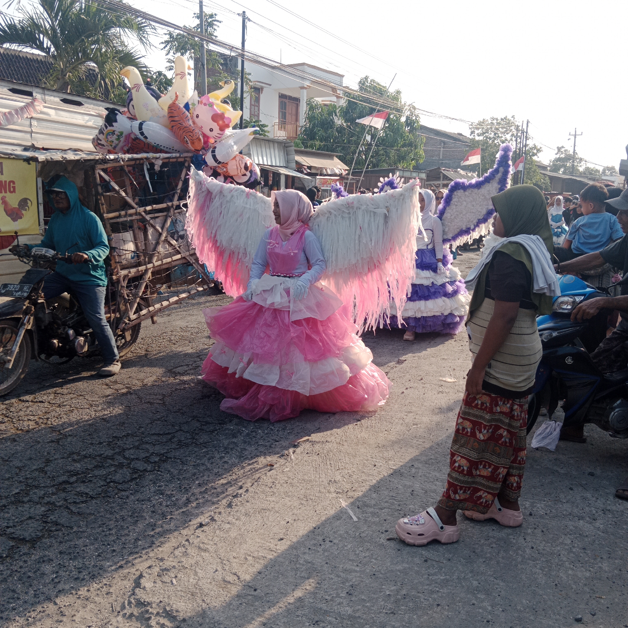 The image depicts a street scene in Tegalrejo, Indonesia, likely during a parade or festival. In the foreground, a young person wearing a pink dress and large white feathered wings stands prominently. They are also wearing a pink headscarf and white gloves, and their dress has multiple layers of pink and white ruffles. To their right, another person in a green headscarf and patterned pants is partially visible, facing away from the camera. Behind the main subject, other individuals are present, including a child in a blue shirt and adults. A cart filled with colorful balloons, including a Hello Kitty balloon, a tiger-striped balloon, and various animal-shaped balloons, is positioned behind a stall. A person in a blue hooded jacket is riding a motorcycle to the left of the frame, pulling a wooden cart. The street is paved with asphalt and shows signs of wear and tear. In the background, residential buildings with tiled roofs and trees are visible. Indonesian flags are displayed on poles. The lighting suggests it is either early morning or late afternoon, with long shadows cast across the ground.