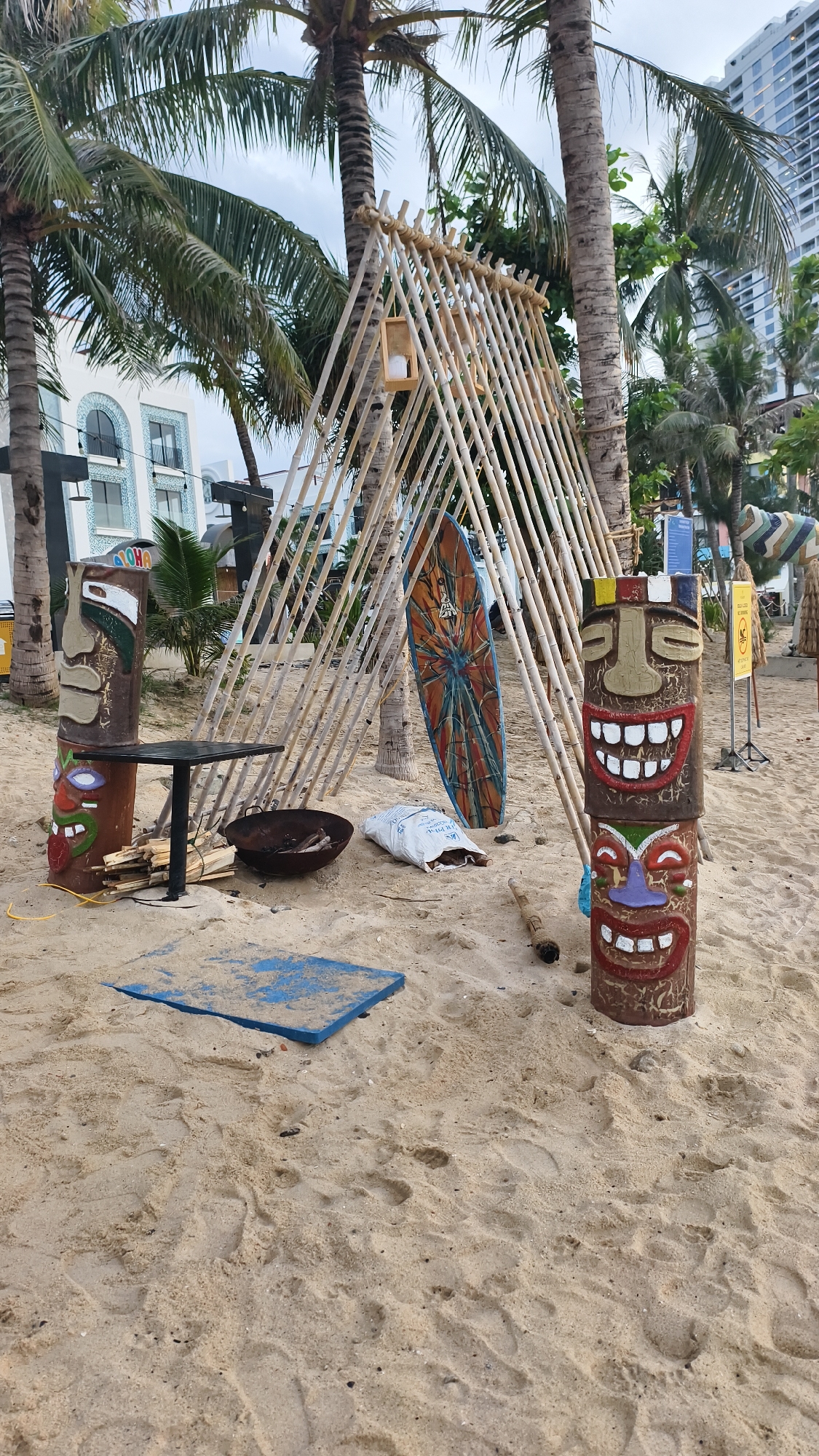 The image shows a beach scene in Nha Trang, Vietnam, featuring a decorative tiki-style setup. Two carved tiki statues stand on the sand, one with a "Aloha" inscription. A surfboard with a colorful, abstract design leans against a structure made of bamboo poles. In front of the bamboo structure, there is a small black table with a shallow metal bowl containing what appears to be ash and remnants of burnt material. Next to the bowl is a white bag, possibly filled with sand. A blue square object is placed on the sand in the foreground. Palm trees are visible throughout the scene, with buildings in the background suggesting an urban coastal environment. The sky is overcast. The scene appears to be set up for a beachside event or decoration.