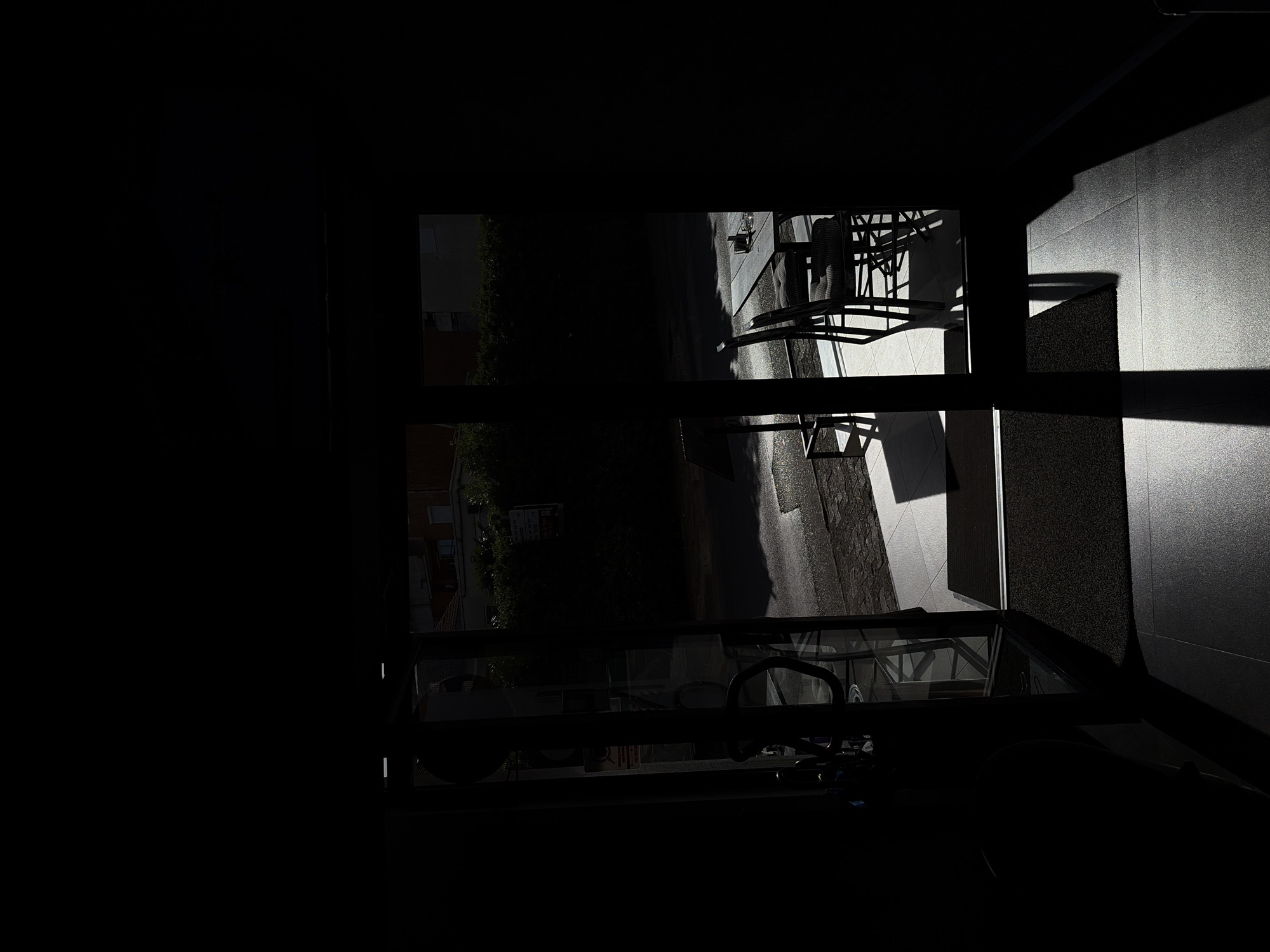 From inside a darkened interior, a window reveals a partial view of an external area. The scene shows parts of buildings, trees, a paved walkway, and metal structures with angular designs. The play of light and shadow is pronounced, with strong contrast. The location is possibly in Budva, Montenegro.