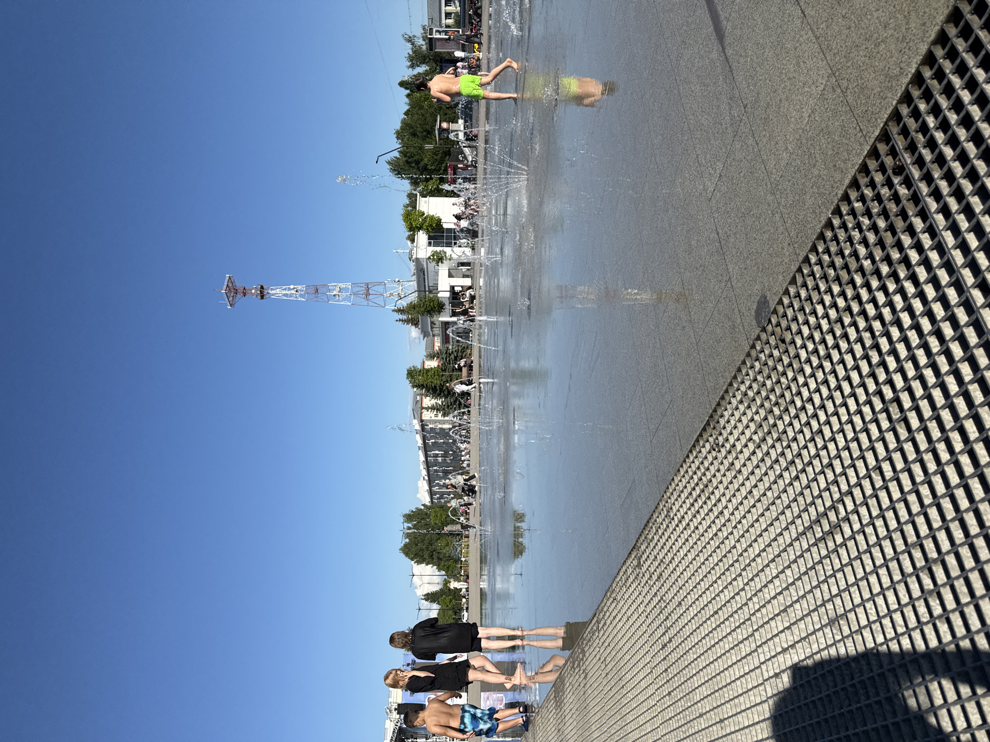 The image depicts a modern, open public space with a large, shallow fountain on a sunny day in Izhevsk, Russia. Several children and adults are present, some playing in the water. In the foreground, a metal grate runs along the edge of the fountain area, reflecting the bright sky. To the left, three figures, two children and one young adult, are standing near the edge of the fountain. One child is wearing blue and white patterned swim trunks, while the other two are dressed in dark, possibly black, clothing. Further into the fountain, a young person wearing bright green swim trunks is walking through the spraying water.
In the background, there are low-rise buildings, trees, and a tall, red and white telecommunications tower. Other people are visible in the distance, some sitting or walking. The overall scene suggests a recreational area in a city, possibly a city square or park, designed for public enjoyment during warm weather. The sky is a clear, vibrant blue, indicating a cloudless day. The architecture and the presence of the telecommunications tower suggest an urban environment within Izhevsk.