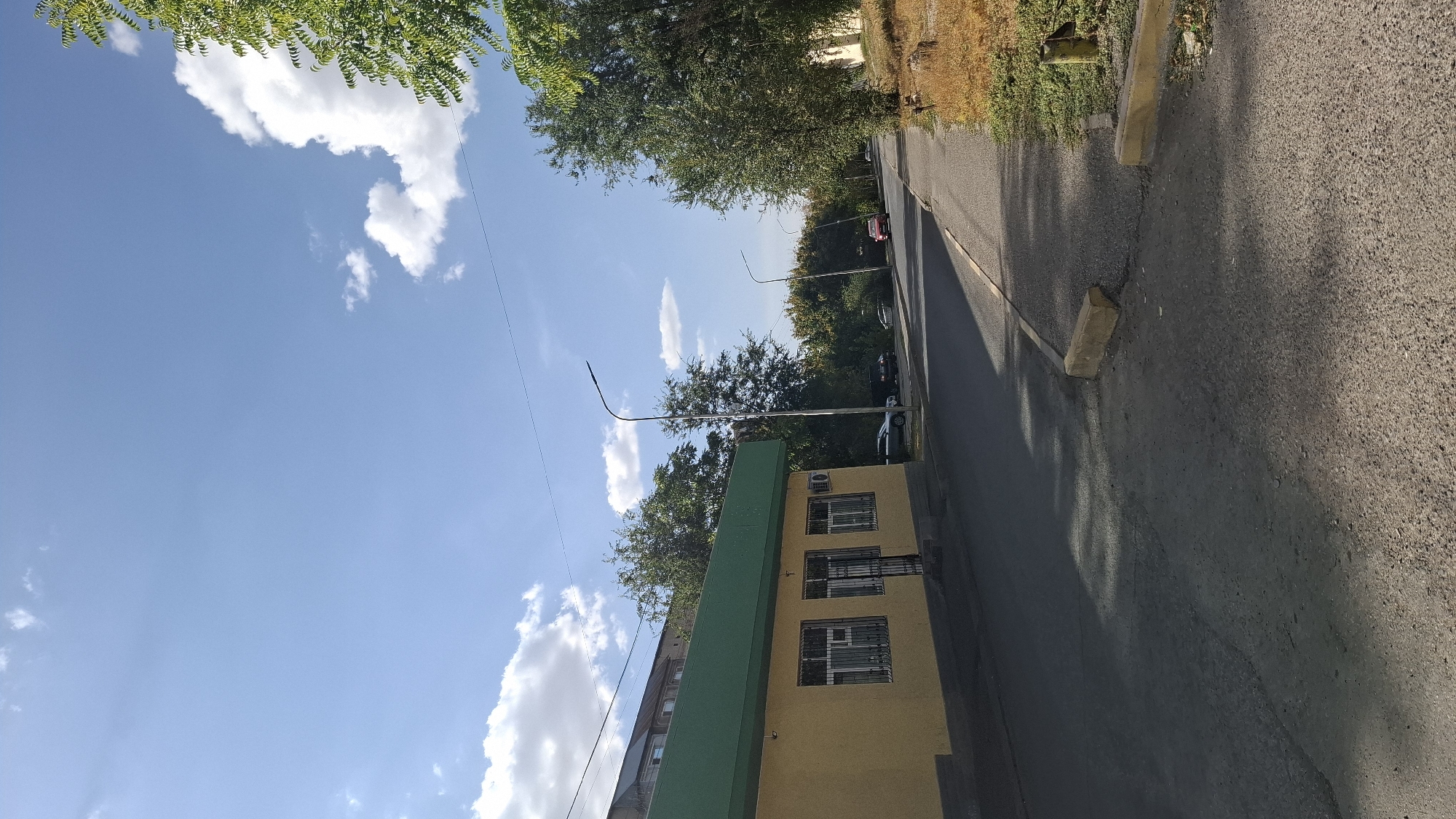 The image depicts a street scene in the city of Shymkent, Kazakhstan. On the left, a single-story building with a yellow facade and a green overhang is visible. The building has barred windows and an air conditioning unit. Adjacent to the building is a paved road. Further down the road, several cars are parked and in motion, including a red sedan. Tall trees line the right side of the road, some with dense green foliage and others with drier, autumnal colors. The sky above is blue with scattered white clouds, and sunlight casts shadows on the road. The scene appears to be an outdoor environment within a residential or commercial area of the city.