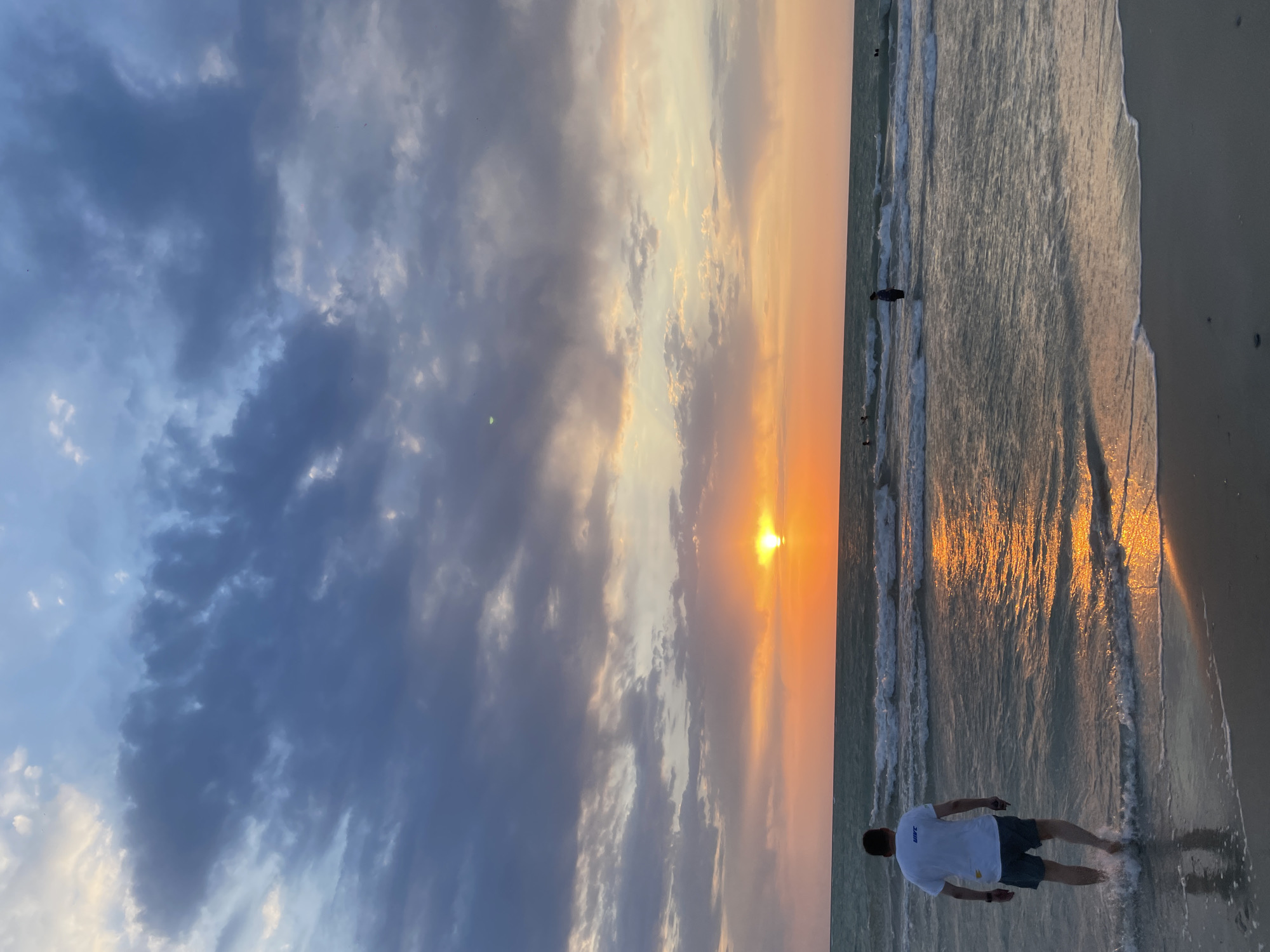 An individual wearing a white t-shirt and dark shorts stands in the shallow water at the edge of a beach, facing away from the camera towards a sunset over the ocean. The sun, a bright orange orb, is low on the horizon, casting a golden reflection on the water. Waves gently break on the shore, and other people are visible in the distance in the water. The sky is filled with dramatic clouds, with shades of blue, gray, and orange from the sunset. The sand is wet where the waves have receded. The scene is a serene beach sunset.