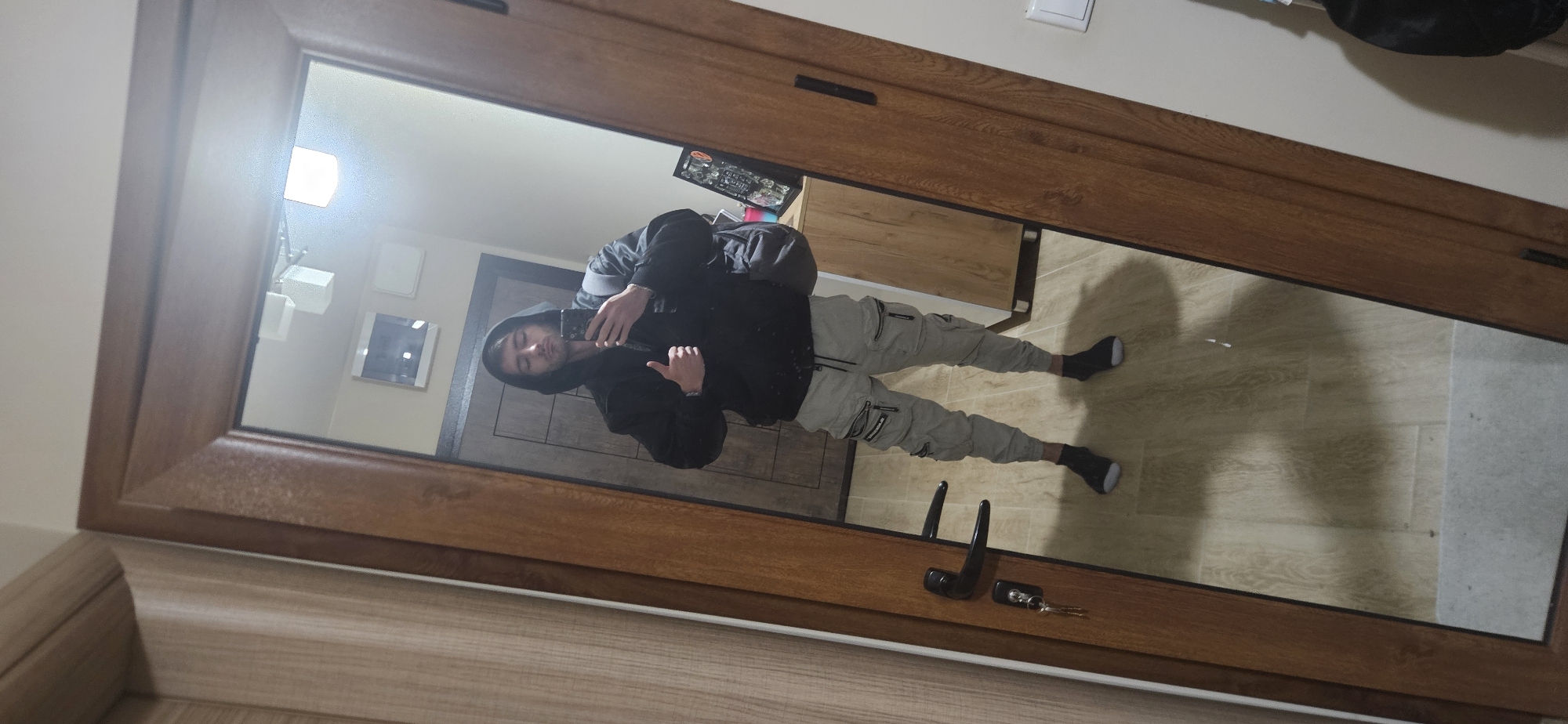 The image is a reflection in a wooden-framed mirror, showing a person taking a selfie. The person is wearing a black hooded jacket and light-colored cargo pants with black shoes. They are holding a phone in their hand. The background behind the person appears to be a hallway with a wooden door and beige walls. Visible in the reflection is a lighting fixture, decorative wall squares, a wooden cabinet, and possibly a jewelry box. On the mirror frame, near the bottom, is a door handle and a set of keys. The floor appears to be tiled in a light brown color. The location is unknown to me, and I can not determine if this is in the city of Plovdiv, Bulgaria.