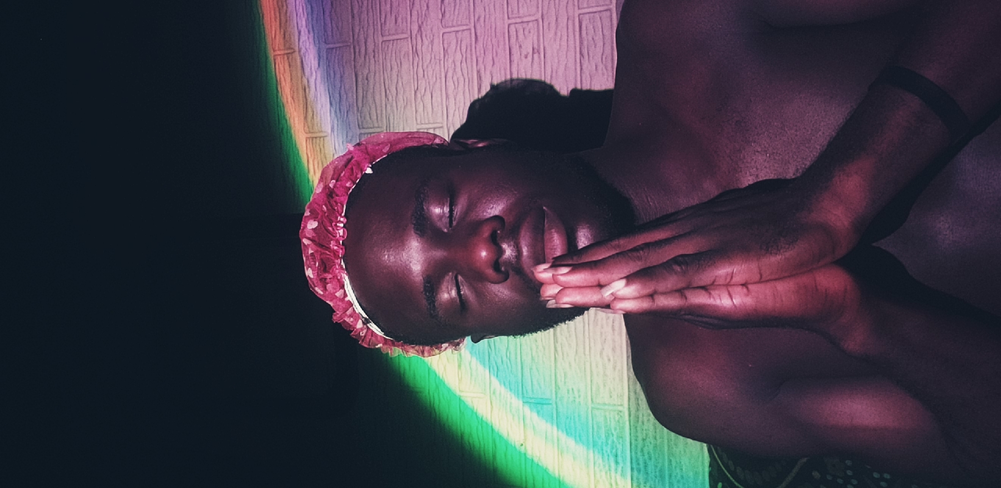 A dark-skinned man wearing a pink floral shower cap lays with closed eyes, hands clasped near his face. He is shirtless and has a black wristband on his right arm. A rainbow light projects onto a textured wall behind him. The location is not specified in the image and cannot be ascertained from the visual information provided. Therefore, no information about the location of Narayi, Nigeria, is included in the description.