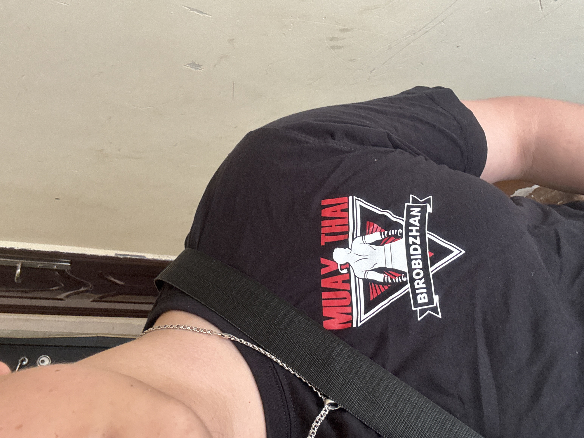 The image shows a person wearing a black t-shirt with a print that reads "MUAY THAI" vertically in red lettering and "BIROBIDZHAN" horizontally in white lettering within a banner surrounding a triangle with a figure inside. A black strap is visible across the t-shirt, and a silver chain necklace is around the person's neck. The background appears to be a plain, light-colored wall. The person's partial arm and shoulder are visible. The location is Unknown, Unknown.