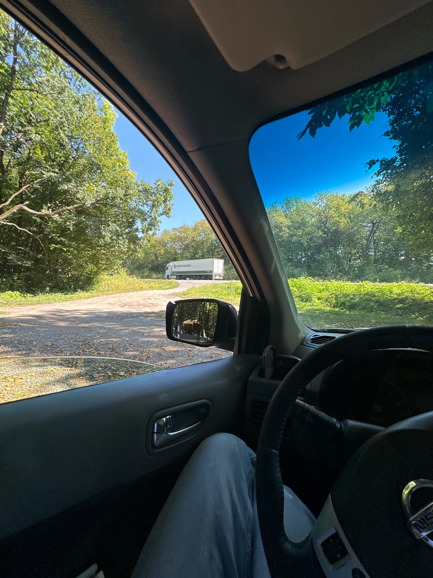 The image is taken from inside a car, looking out the driver's side window towards a dirt road that curves into a wooded area. A white semi-trailer truck with green lettering is visible on the road in the distance. The sky is clear blue. The interior of the car shows a portion of the steering wheel, the driver's leg in blue jeans, and the car door with a silver handle. The side mirror reflects a wooded area. The scene appears to be in a rural or semi-rural environment. The provided context places the scene in Shatava, Ukraine.