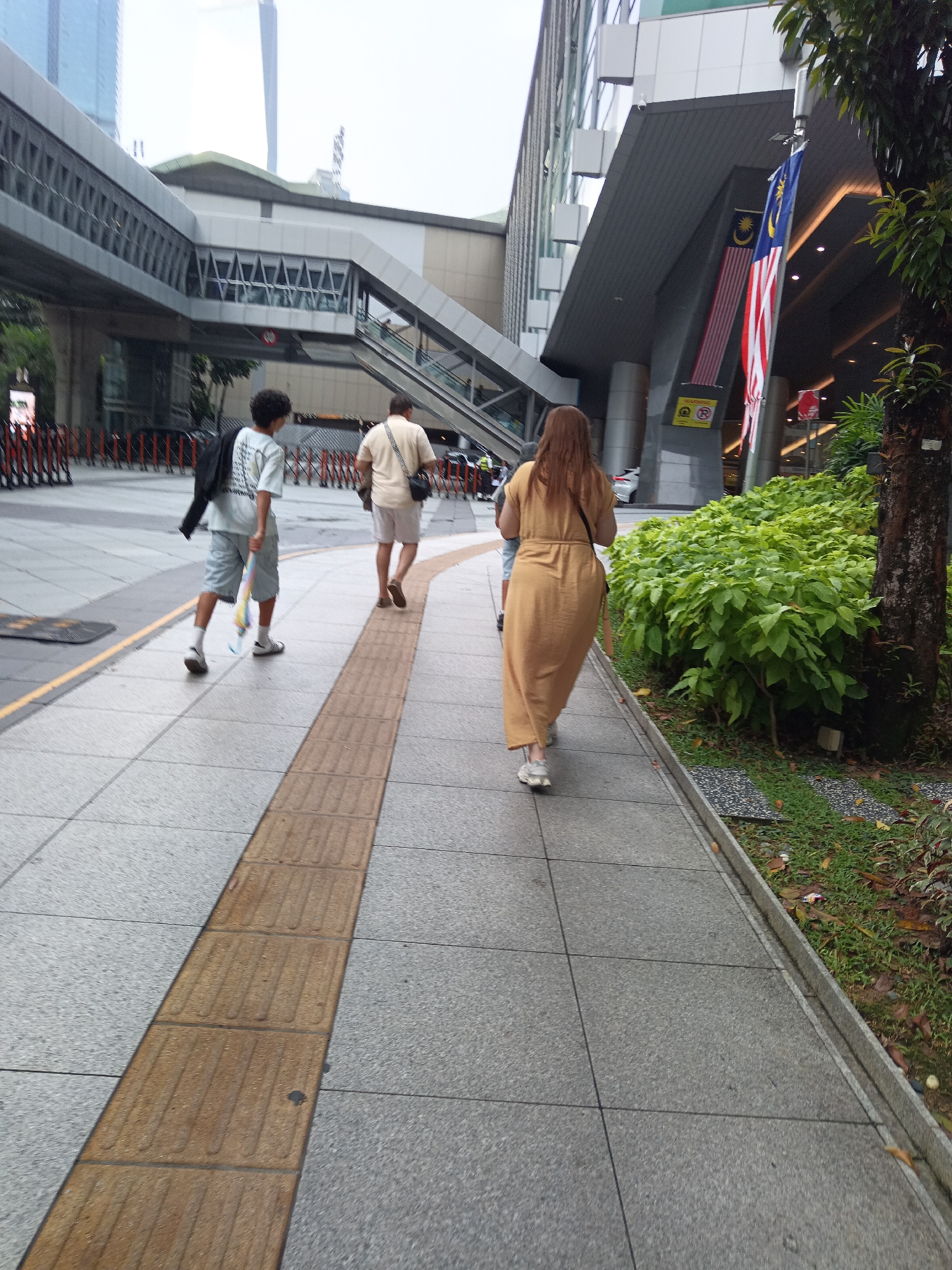 The image depicts a street scene in Kuala Lumpur, Malaysia. A wide, paved sidewalk runs diagonally across the frame, with a textured tactile paving strip bordering the left side. Several individuals are walking along the sidewalk. In the foreground, a person wearing a light-colored t-shirt and light blue shorts, carrying a black jacket over their left shoulder and a colorful object in their right hand, walks away from the viewer. Behind them, a person in a light beige polo shirt and light-colored shorts walks in the same direction, carrying a dark bag over their right shoulder. Further ahead, a person in a long, tan-colored dress walks away from the viewer.
In the background, a modern building complex with elevated walkways and escalators is visible. Malaysian flags are displayed on the building's facade. Green foliage and trees line the edge of the sidewalk, and various signs are present on the building. The ground is paved with square tiles. The overall lighting suggests an overcast day.