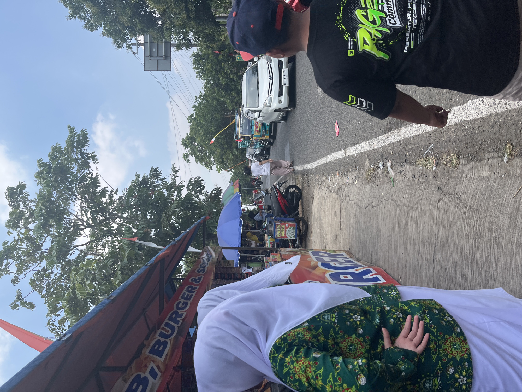 The image depicts a street scene in Glagah Kulon, Indonesia, likely on a sunny day with a partly cloudy sky. In the foreground, a person wearing a black t-shirt with a green graphic design and a blue baseball cap is facing away from the camera, observing the road. To the left, partially visible, are two individuals wearing hijabs, one with a white hijab over a green patterned top and a white skirt, and another with a white hijab. They appear to be standing near a food stall with a red and blue awning. The stall has a banner that reads "BURGER & SOON" and has orange and yellow branding. Several other food vendors with umbrellas and motorcycles are also visible further down the street.
The street itself is paved and marked with a white line. A line of vehicles is proceeding down the road, including a white car with license plate H 1613 YM, a green and blue truck, and other cars and motorcycles. Trees line the sides of the road, and there are overhead wires and a traffic sign in the background. The overall impression is of a busy roadside market or gathering, possibly near a public area, with economic activity taking place alongside traffic. There is some litter visible along the edge of the road.