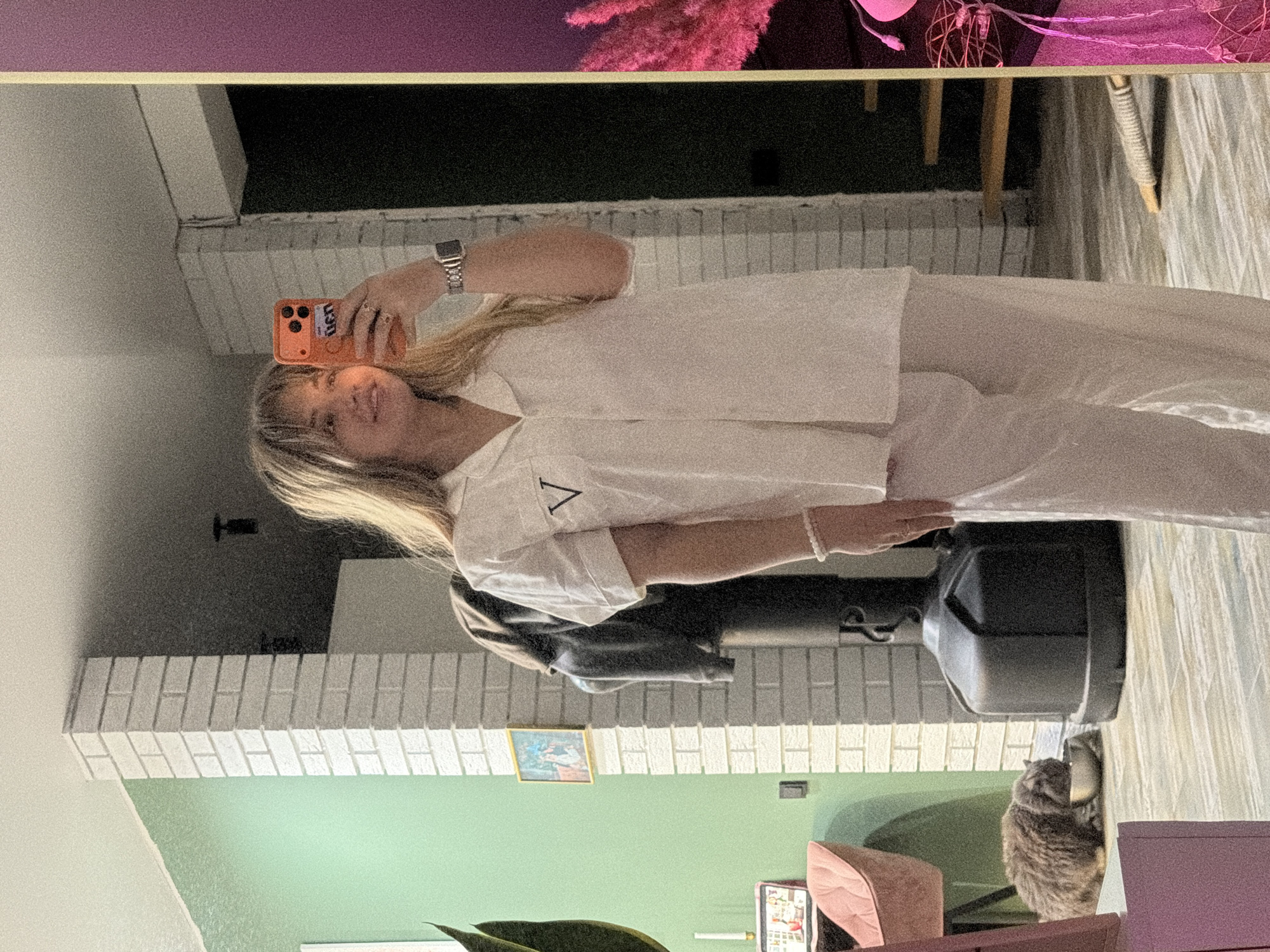 The image shows a woman reflected in a mirror. She is holding a phone, taking a selfie. She is wearing white pajamas with a "V" on the shirt. The background shows a room with a white brick fireplace, a green wall, a picture on the wall, and a cat. The image includes decor, a speaker, and part of a plant. The location is Большая Шильна, Russia.