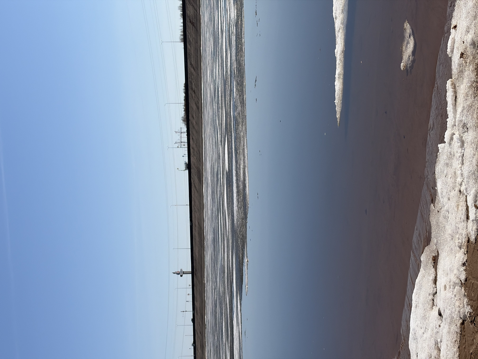 This image, taken in Noviy, Russia, captures a vast, clear landscape under a bright, cloudless blue sky on a sunny day, likely during late morning or early afternoon. The scene is dominated by a large body of water, possibly a reservoir or a wide river, indicating the early stages of spring with significant ice melt.
Across the mid-ground, the water's surface features extensive stretches of thin, breaking ice, which brightly reflect the sun in shimmering streaks. The open water portions are dark blue, mirroring the sky above. Along the distant horizon, a long, flat dark strip of land, possibly an embankment or a distant treeline of bare trees, is visible. Spanning this horizon are numerous utility poles carrying power lines, and several streetlights, suggesting infrastructure alongside the natural environment. A tall, slender white tower, possibly a water tower or a communications tower, stands prominently in the distance against the blue sky, with a smaller, darker structure nearby.
In the foreground, along the right edge of the water, patches of melting, slushy snow and ice, mixed with earth, define the immediate bank. No people are visible, and no specific activity is occurring. The image conveys a sense of peaceful transition from winter to spring, highlighting the interaction between a broad natural environment and distant human-made structures. There is no visible text in the image.