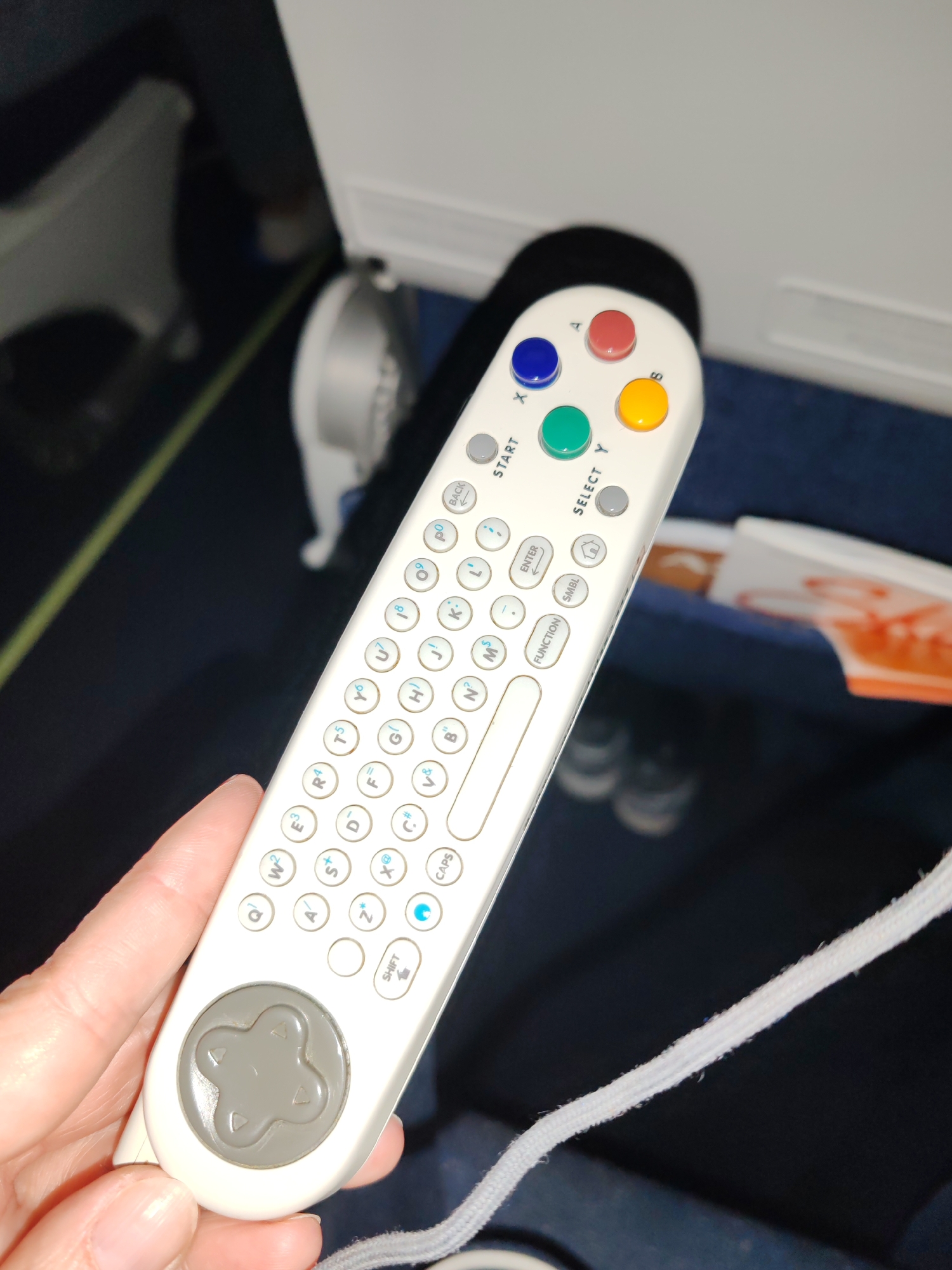 The image captures a close-up of a person's hand holding a white in-flight entertainment remote control inside an airplane cabin. The setting shows a blue carpeted floor and parts of a white airplane interior, including what appears to be a seat structure and possibly a bulkhead or screen housing in the background. A small, blurry white fan-like object is also visible. In the seat pocket to the right, a magazine or papers with orange and white colors can be seen.
The remote control itself is the primary focus. It features a grey directional pad (D-pad) at the bottom. Above it is a mini QWERTY-style keyboard with numerous small, circular white buttons, many of which show multiple characters (letters, numbers, and symbols) such as "Q W²", "E³", "R⁴", "T⁵", "U⁷", "I⁸", "O⁹", "P⁰", along with other letters like A, S, D, F, G, H, J, K, L, Z, X, C, V, B, N, M. Special function buttons include "BACK", "ENTER", "FUNCTION", "SMBL" (symbols), a "HOME" icon, "SHIFT", and "CAPS". At the top of the remote, there are four colored action buttons (blue 'X', red 'A', green 'Y', yellow 'B') and two grey buttons labeled "START" and "SELECT". A white, braided cord extends from the bottom right of the remote, indicating it is tethered.
The activity depicted is the act of holding and potentially operating this remote, likely to control an in-flight entertainment system during a flight. The location context indicates the image is from Saint Petersburg, Russia, but the scene itself is an airplane cabin.